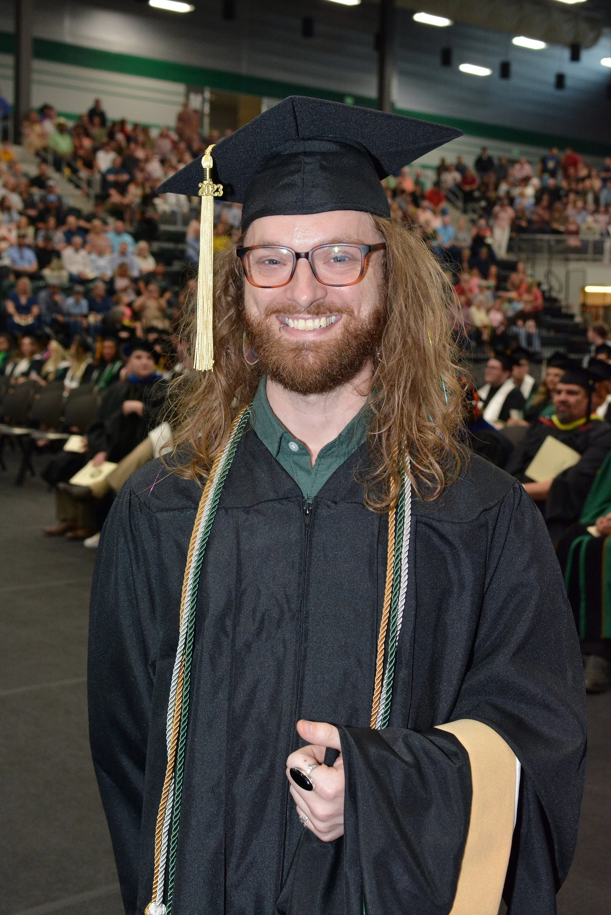 A smiling man with long curly hair, glasses, and a beard in graduation regalia at a graduation ceremony, with an audience of graduates and guests seated in the background.