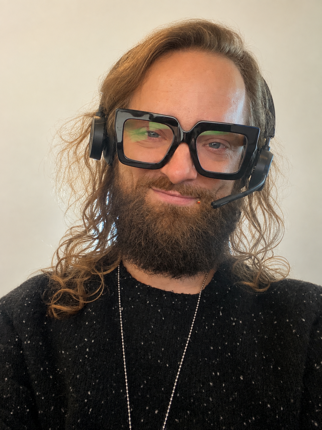A man with long, wavy hair and a beard, wearing oversized black glasses and a headset with a microphone, smiling at the camera.