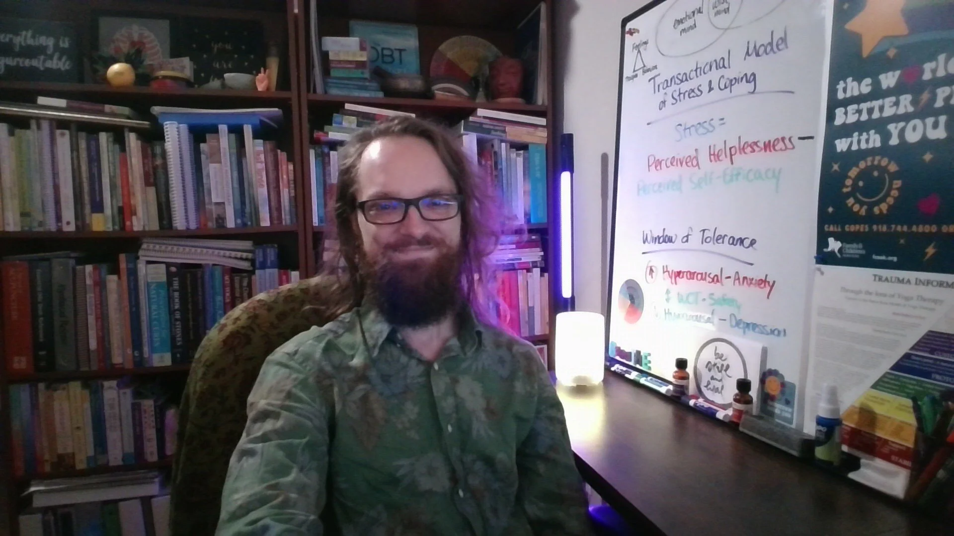A man with glasses and long hair sits in front of a bookshelf filled with books. Behind him, a whiteboard displays notes about stress and psychological concepts, with some colorful decorations and bottles on the table.