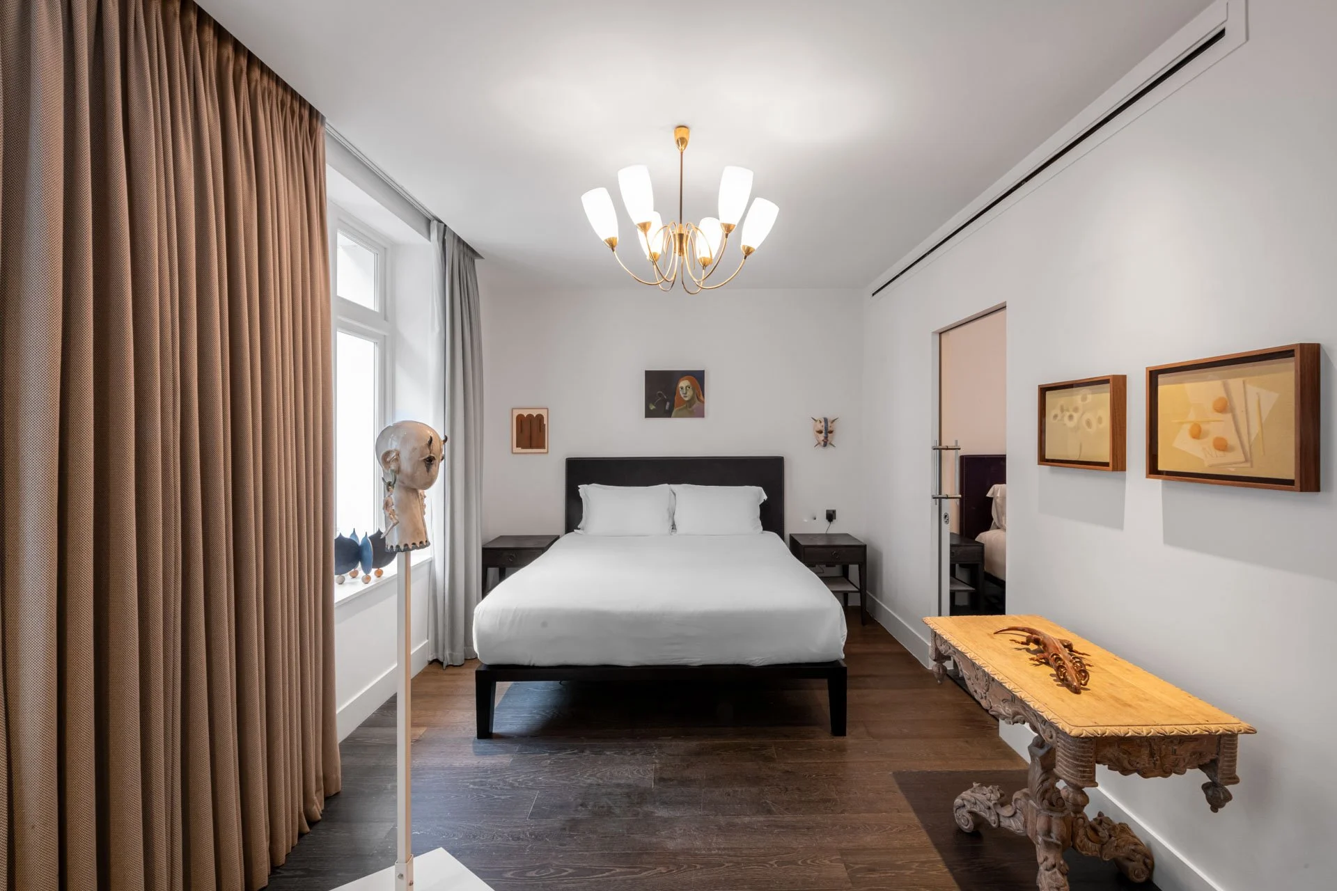 Bright hotel bedroom installation with a made bed, chandelier, heavy   curtain, and framed artworks staged like a lived-in room