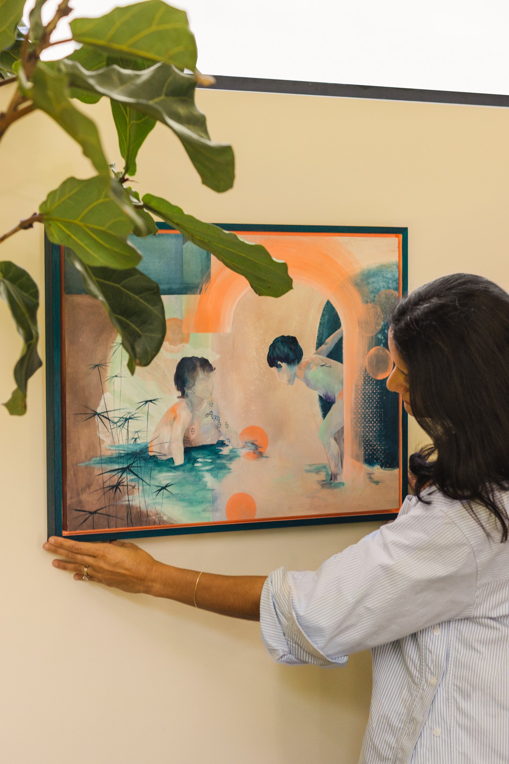 A woman with dark hair and glasses holds a framed painting in front of a wall with a potted plant in the foreground. The painting depicts abstract figures and shapes with warm and cool colors.