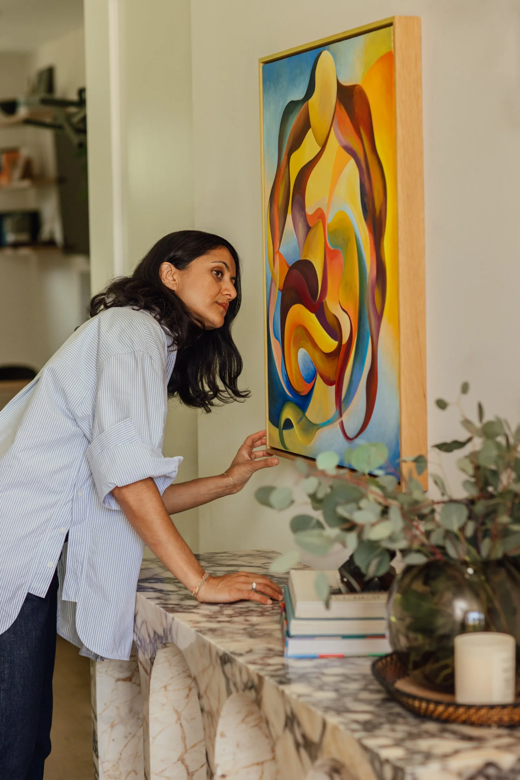 Woman with dark hair in a striped shirt leans over a marble table, viewing an abstract painting on the wall. In the foreground is a potted plant with green leaves and some books.