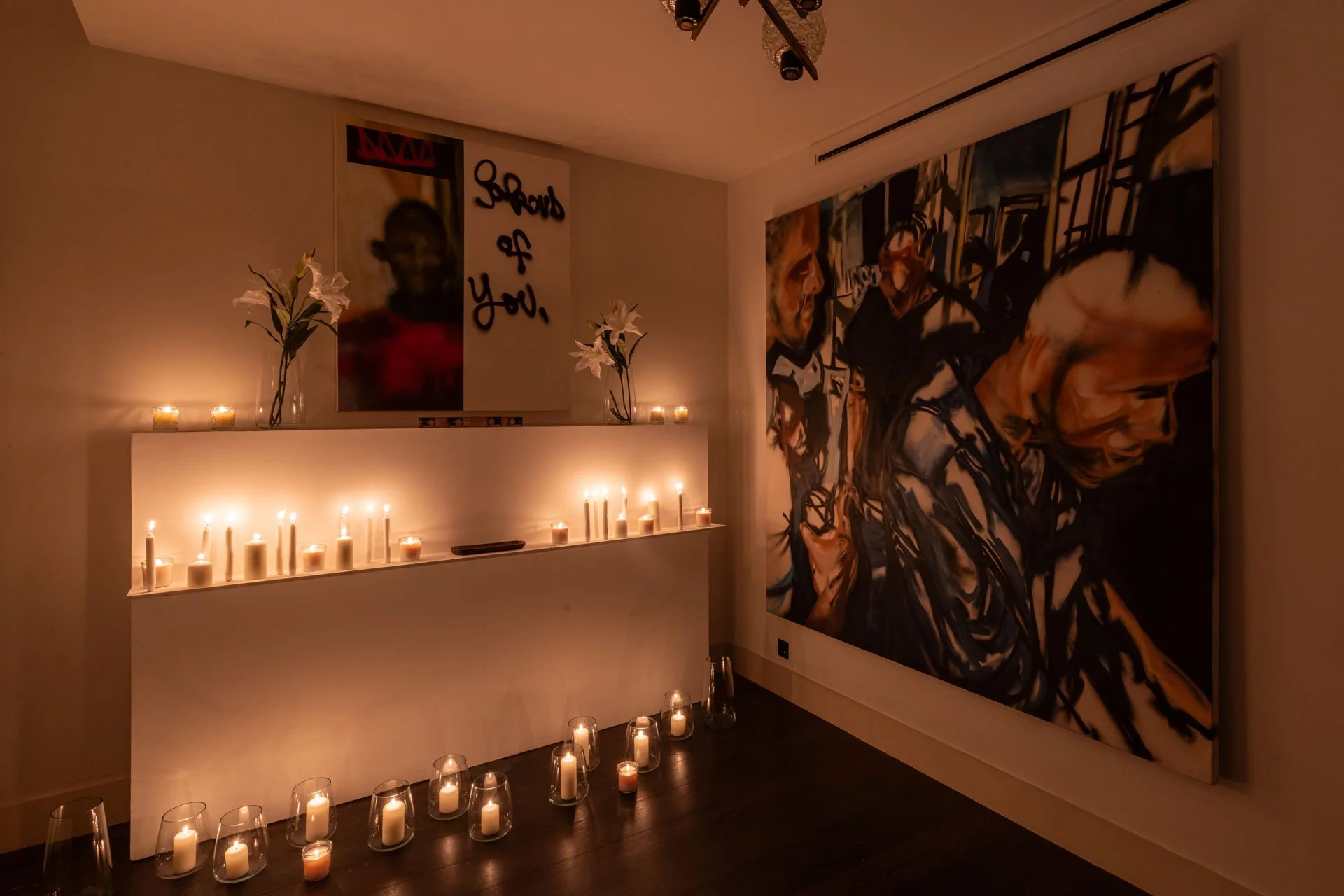 Low-lit room corner with rows of lit candles along the floor and a wall   shelf, creating a warm, devotional atmosphere around the artwork