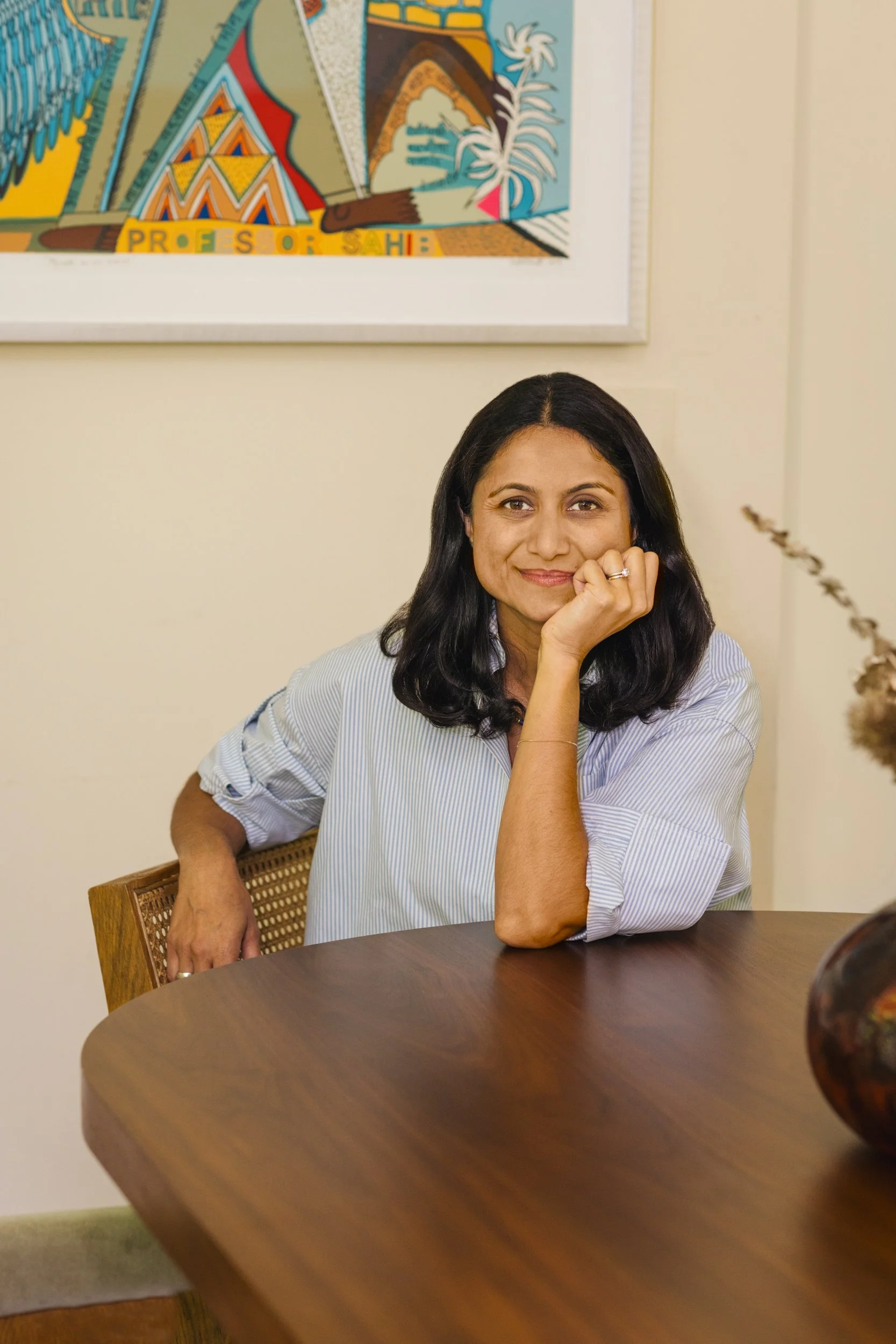 A woman sitting at a wooden table, resting her chin on her hand, smiling at the camera, with artwork on the wall behind her.