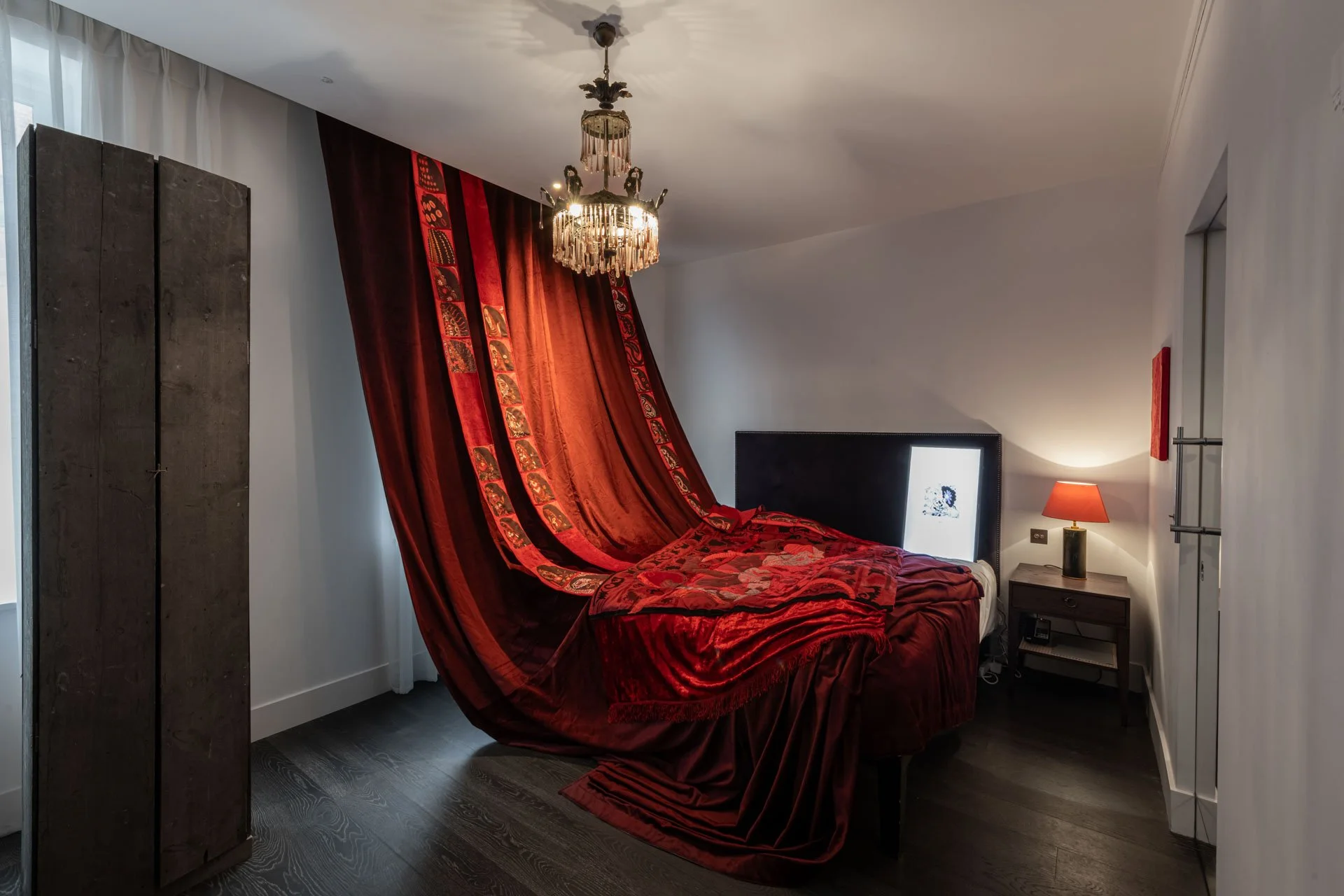 Dim hotel room installation with a dramatic red fabric draped from ceiling   to floor, transforming the space into a sculptural environment