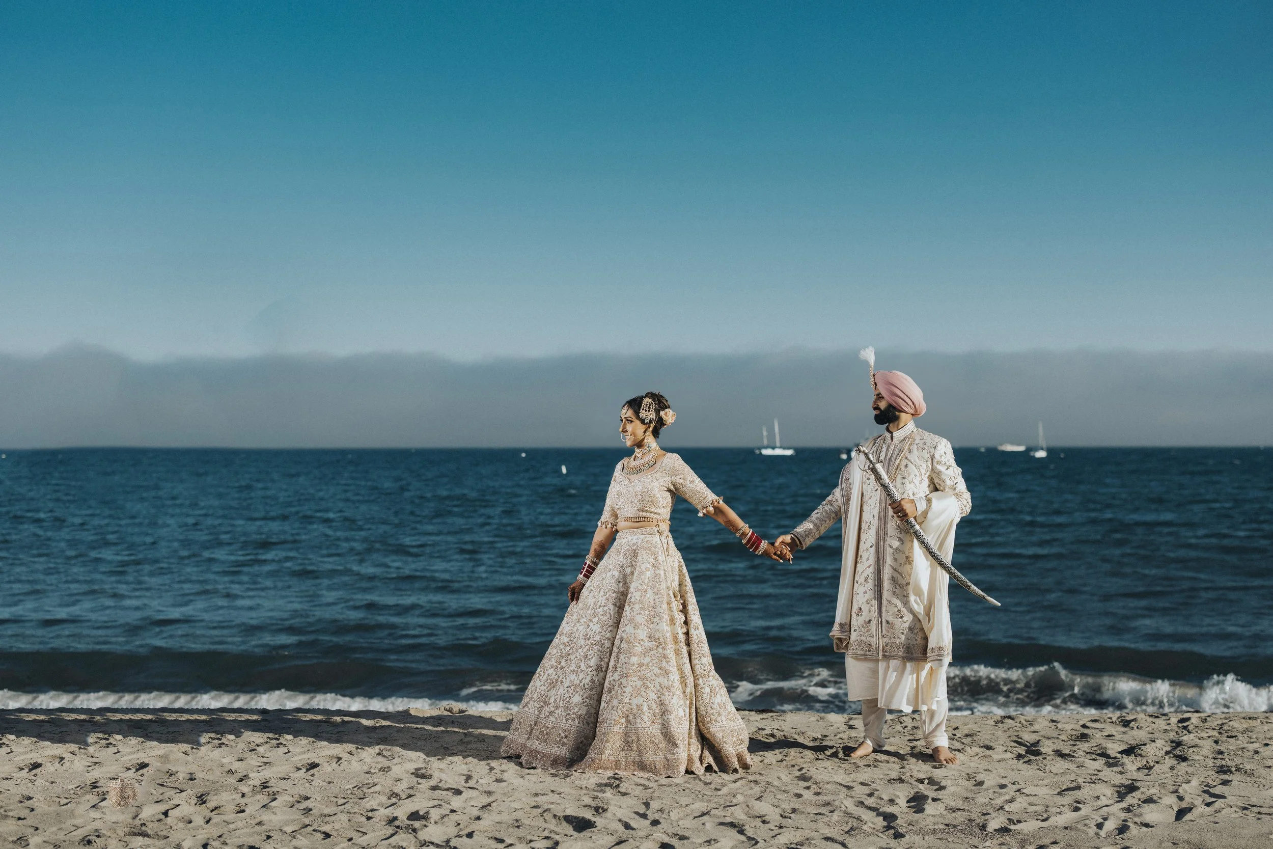 An Indian couple in traditional wedding attire holding hands on a beach, with the ocean and sailboats in the background.