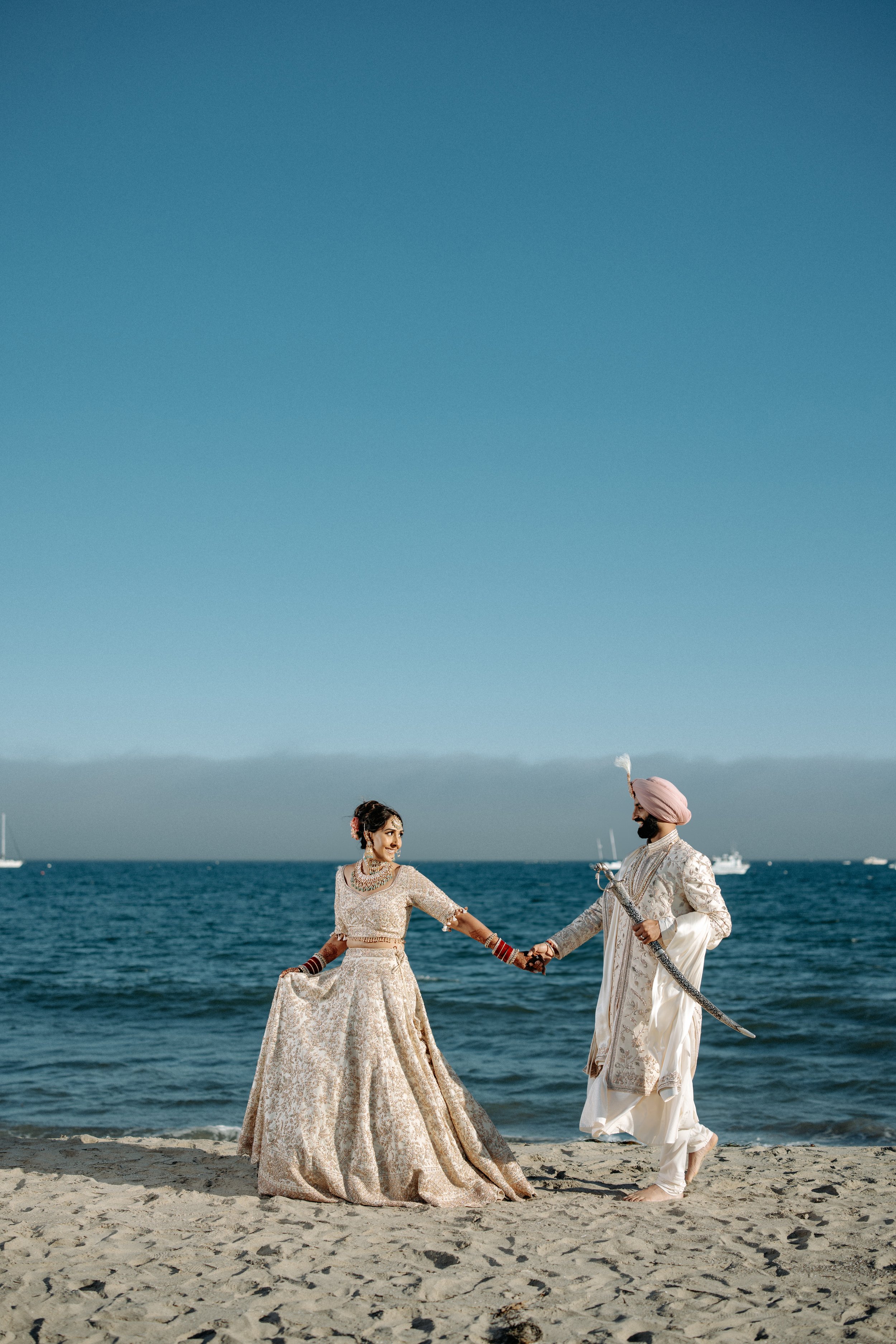 A couple dressed in traditional Indian wedding attire stands on a sandy beach by the ocean holding hands, with a boat visible on the water in the background.