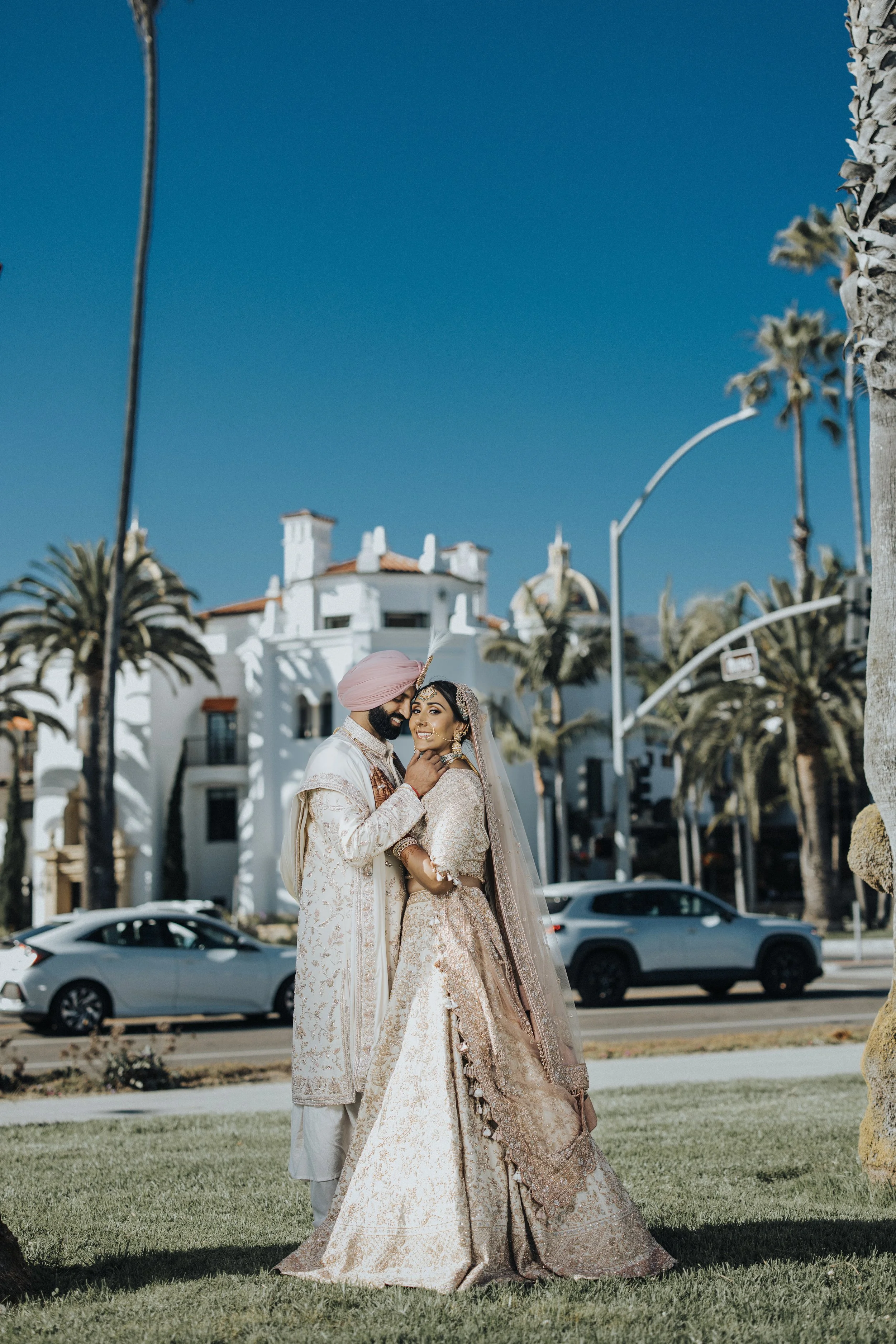 A newlywed couple dressed in traditional Indian wedding attire, standing close together outdoors on a sunny day, with palm trees and a white building in the background.
