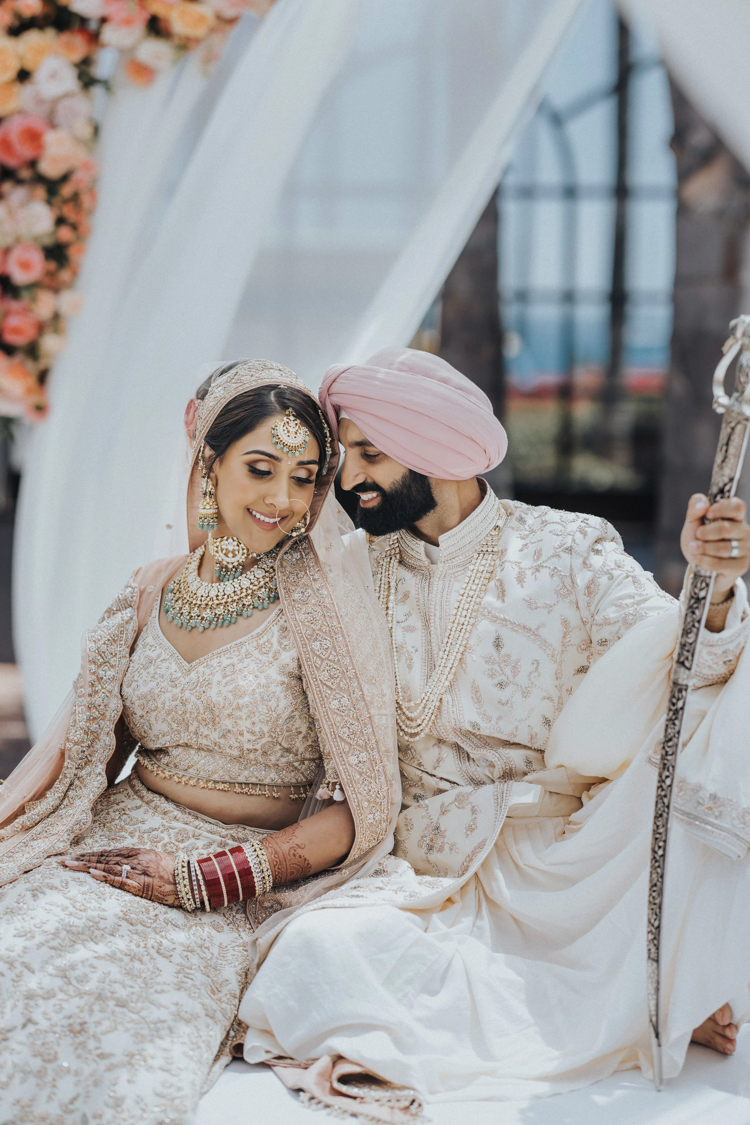 A bride and groom dressed in traditional Indian wedding attire sitting closely together, smiling, with the bride showing henna on her hand and adorned with jewelry, including a necklace, earrings, and a headpiece, and the groom wearing a sherwani and