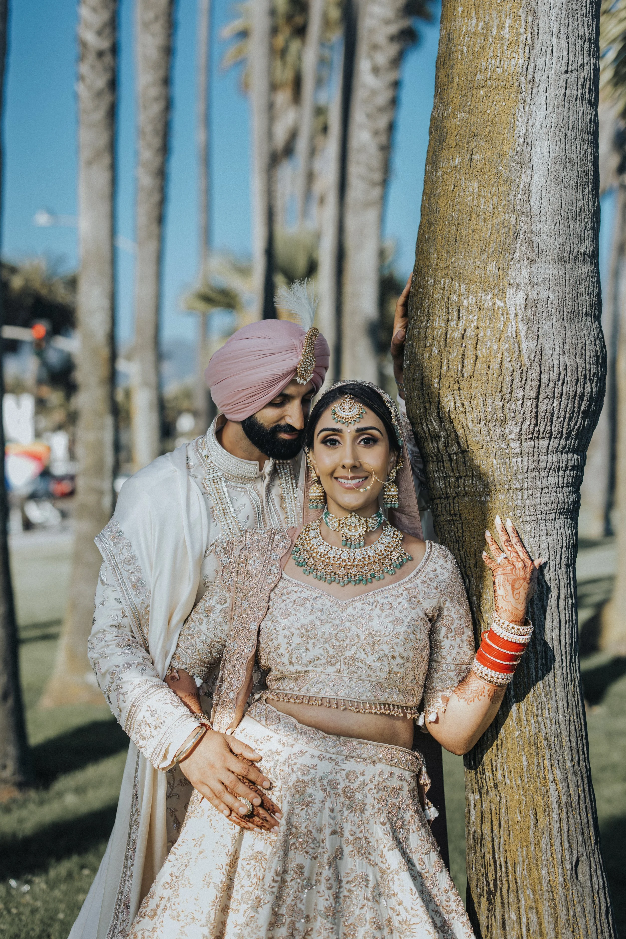 A newlywed Indian couple dressed in traditional wedding attire, standing outdoors next to a palm tree, smiling. The bride is wearing a beige and gold embroidered lehenga with jewelry and henna on her hands. The groom is in a cream sherwani and a pink