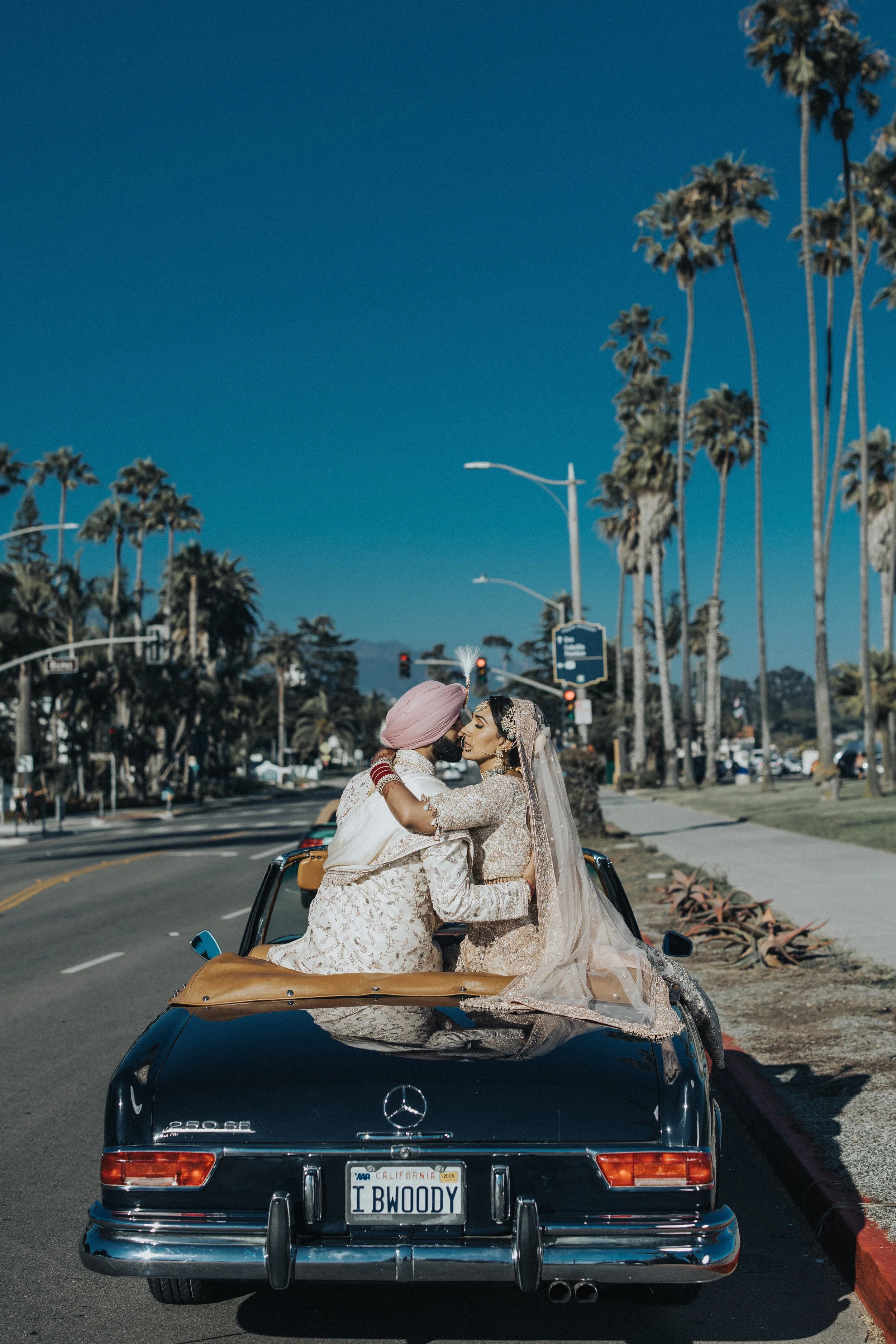 A bride and groom in traditional wedding attire sharing a kiss while sitting on the back of a vintage black Mercedes-Benz convertible with a personalized license plate that reads 'I BWODY', parked on the side of a street lined with palm trees.