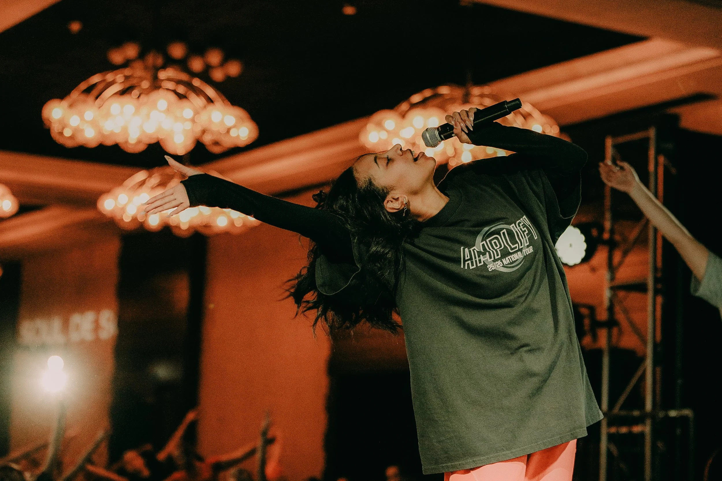 Woman with dark wavy hair, wearing a black T-shirt, speaking or presenting on a stage with a large screen and bright lights or chandeliers overhead.