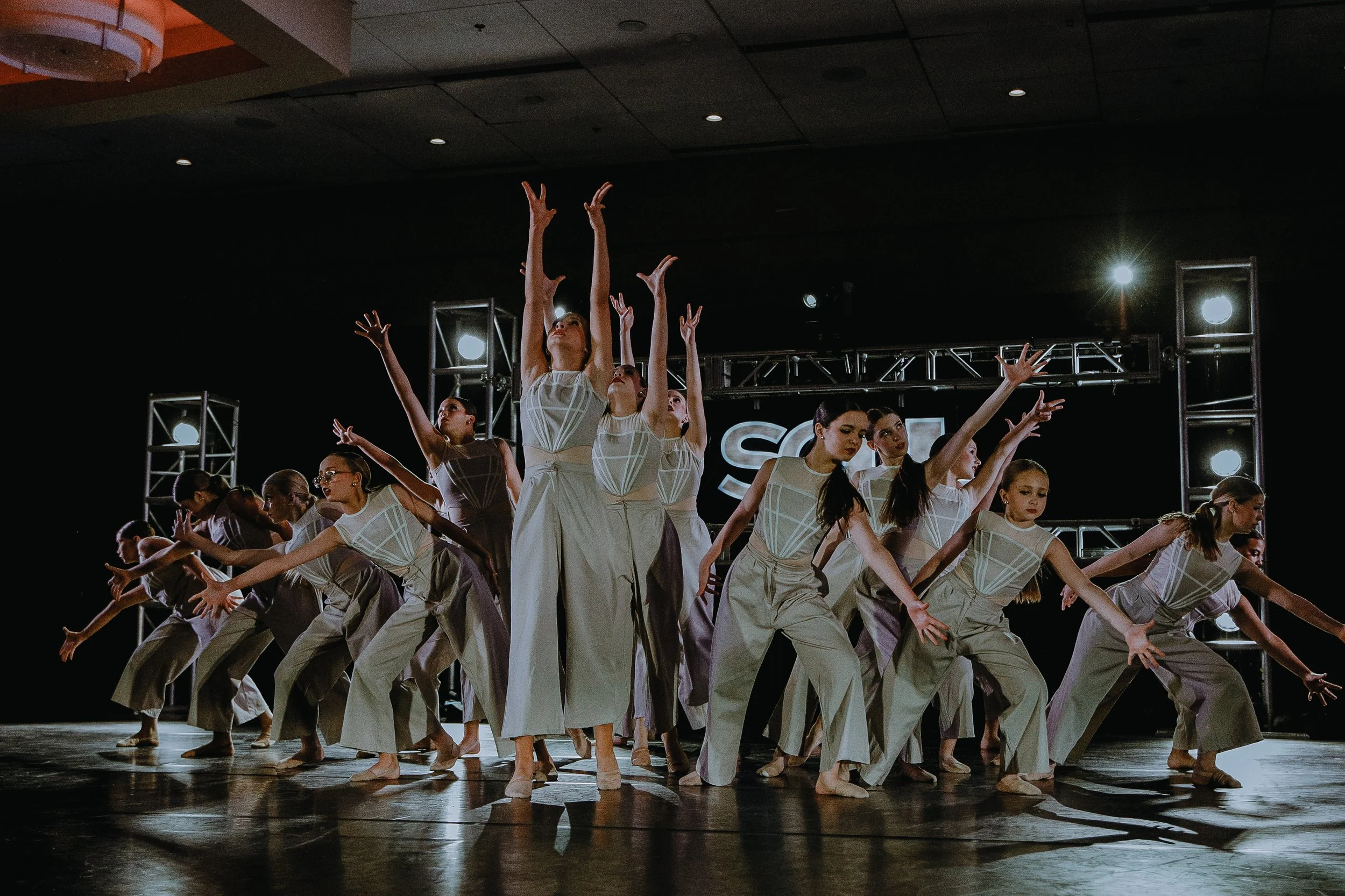 A large group of dancers in beige costumes perform on stage, raising their arms in unison with some bending forward, under stage lighting with a dark background.