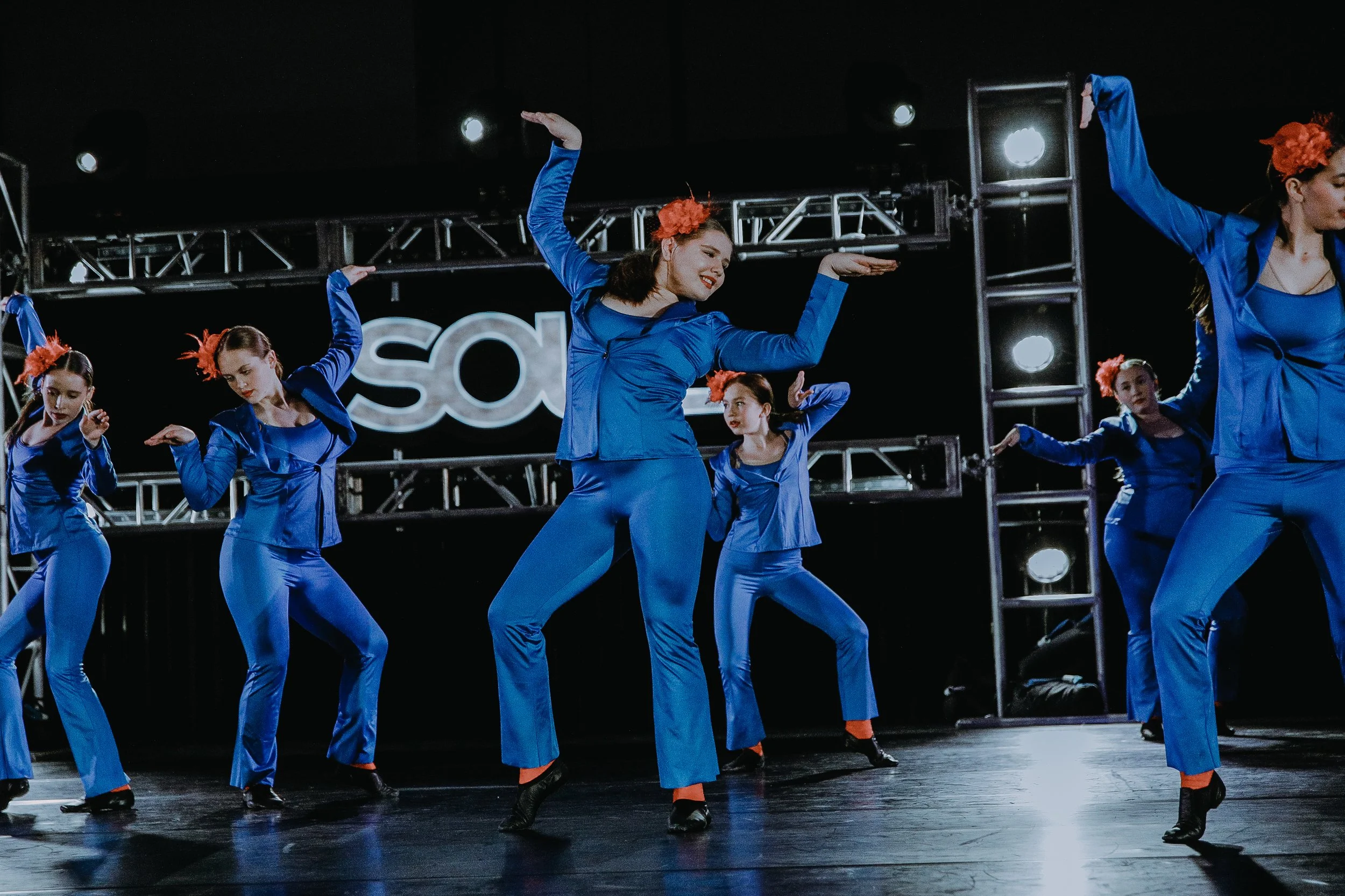 A group of women dressed in vibrant blue costumes performing a choreographed dance on stage with stage lights and a backdrop with the letters 'SO' displayed.
