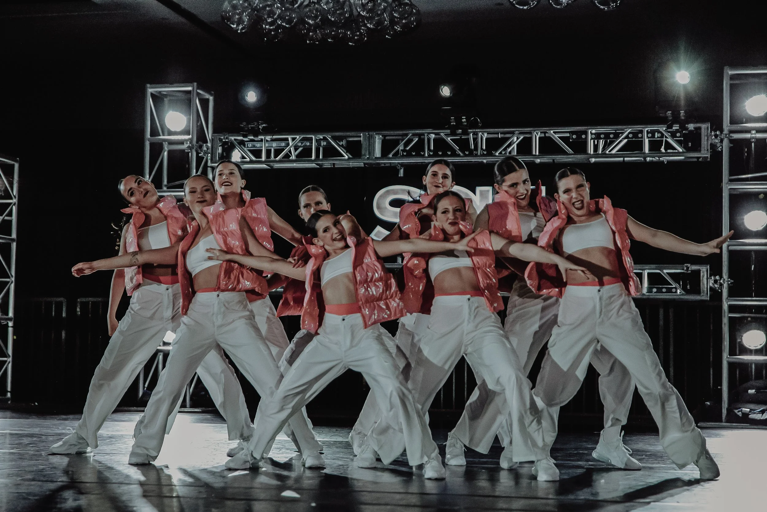 A group of eight female dancers performing on stage with stage lighting and a metal scaffolding structure in the background. They are dressed in white trousers, white tops, and pink shiny vests.