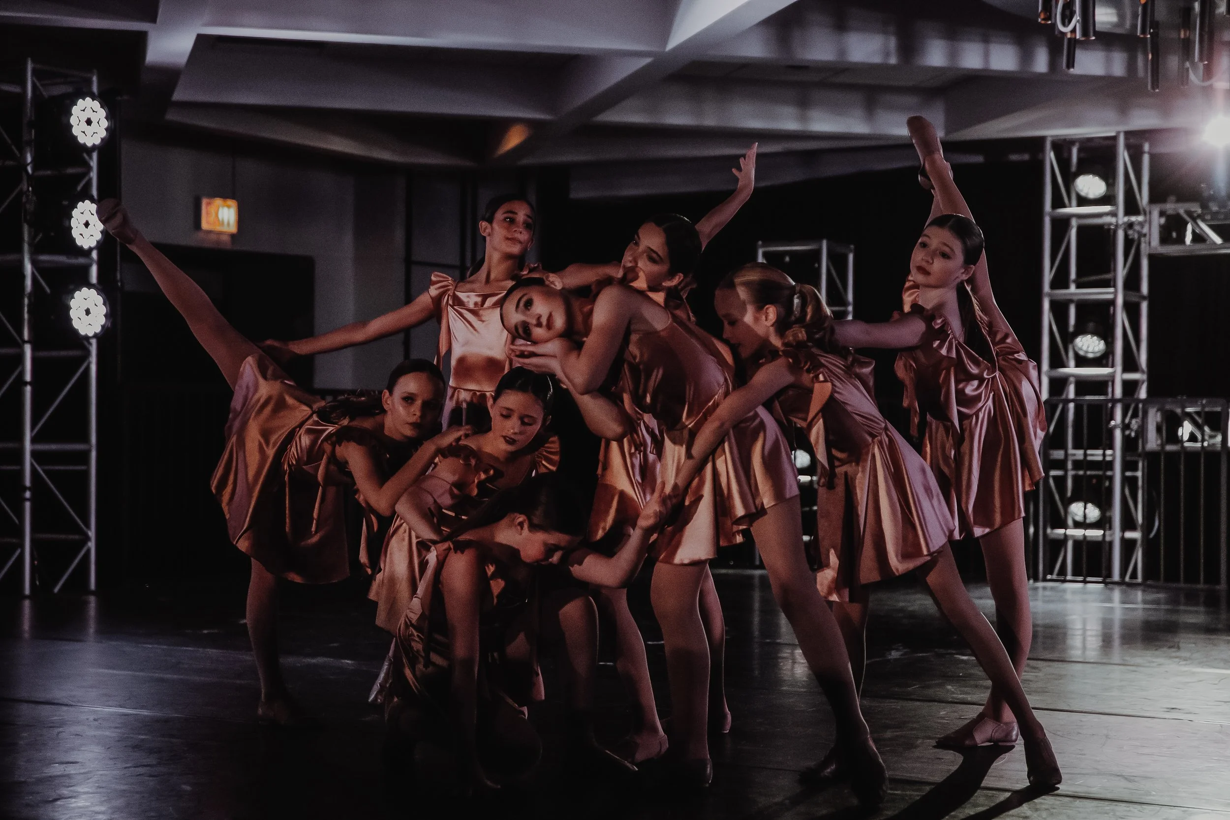 A group of young ballerinas in matching satin dresses perform a dance routine on a stage illuminated with theatrical lighting.