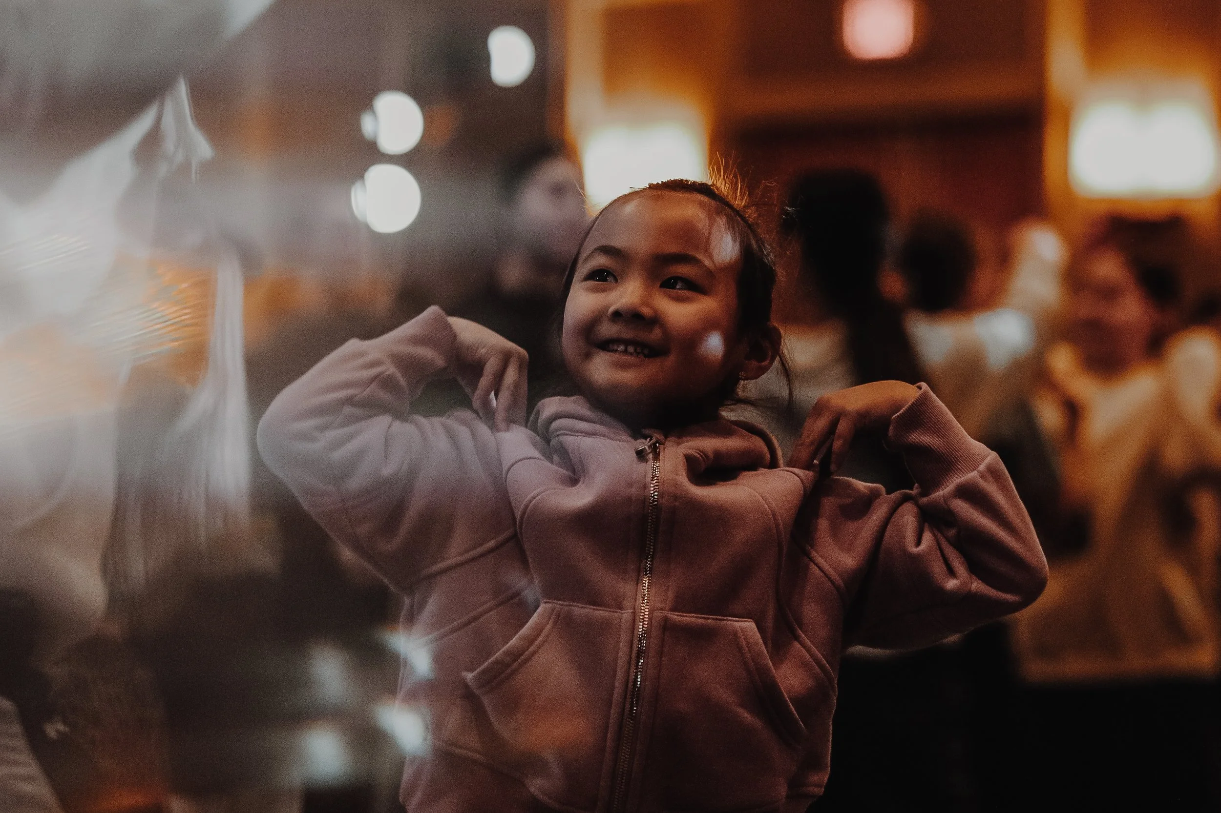 A young girl with a smiling face wearing a light pink hoodie, inside a warm-lit room, with bokeh lights in the background.