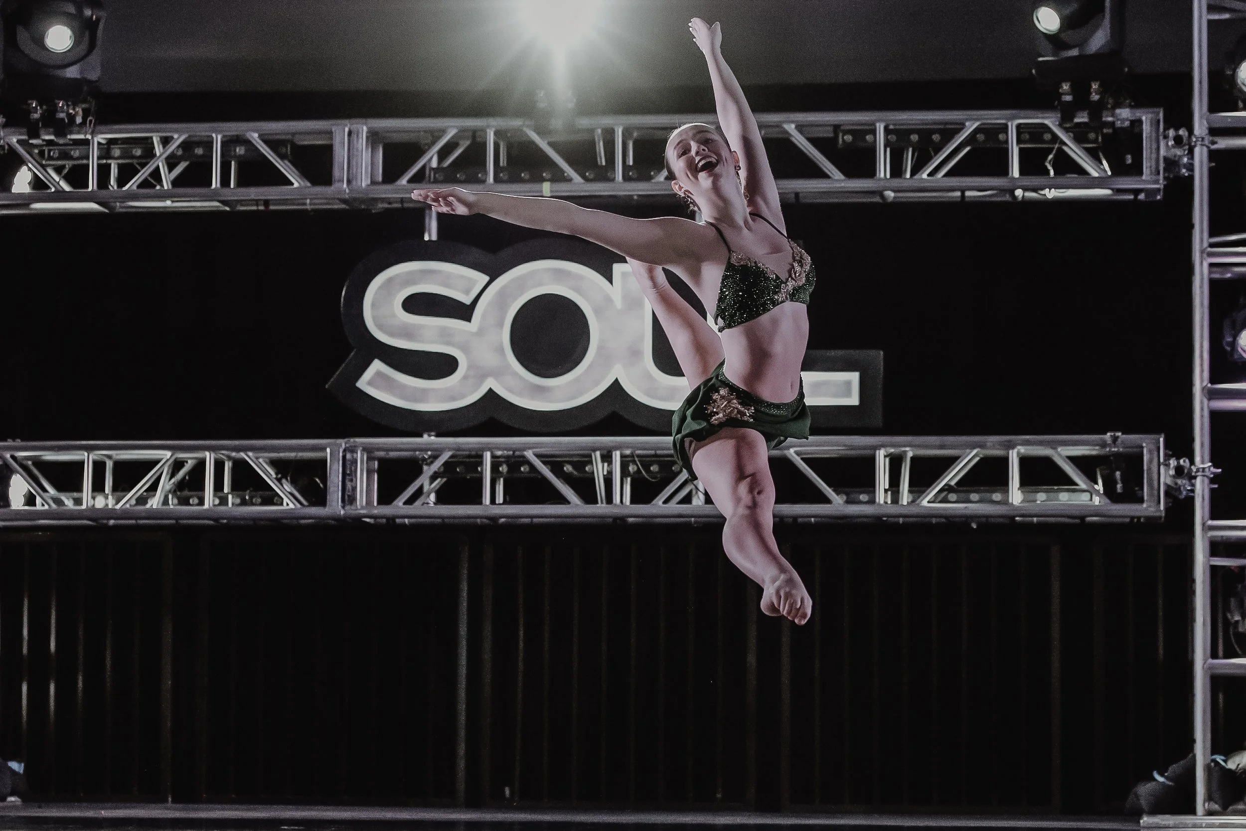 A female dancer performing on stage with her arms raised, wearing a sparkly dance costume, in front of a backdrop that reads 'SOU' with stage lighting above.
