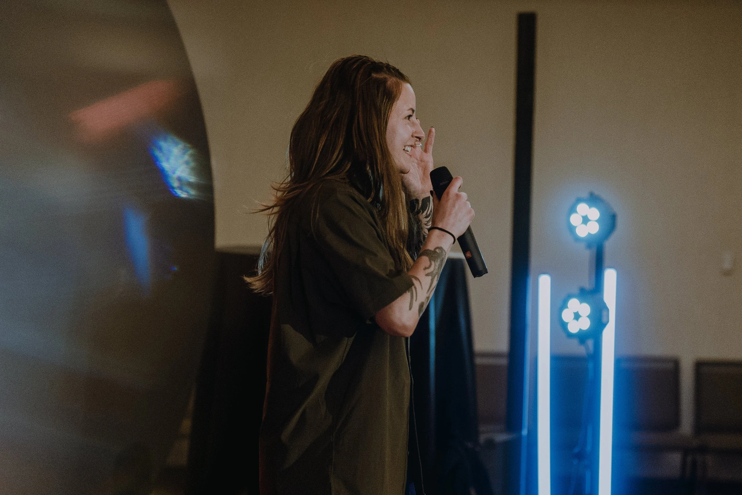 A woman with tattoos on her neck and arms holding a microphone, speaking at an event with a large screen behind her displaying French text.