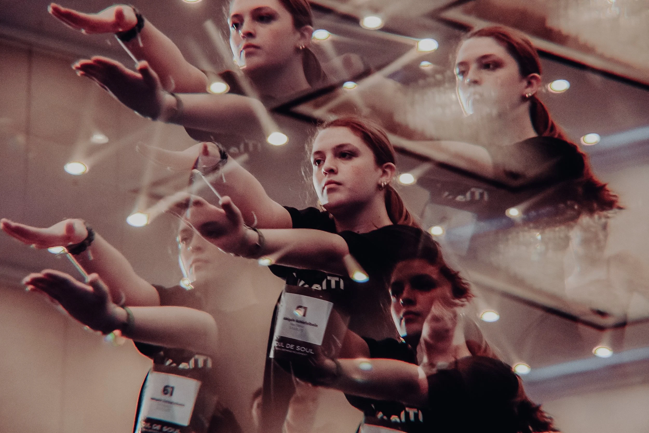 Reflection of a woman athlete with red hair, wearing a black shirt and a race bib, reaching forward with her right hand during a sports event. The reflection is seen on a mirrored surface above her.