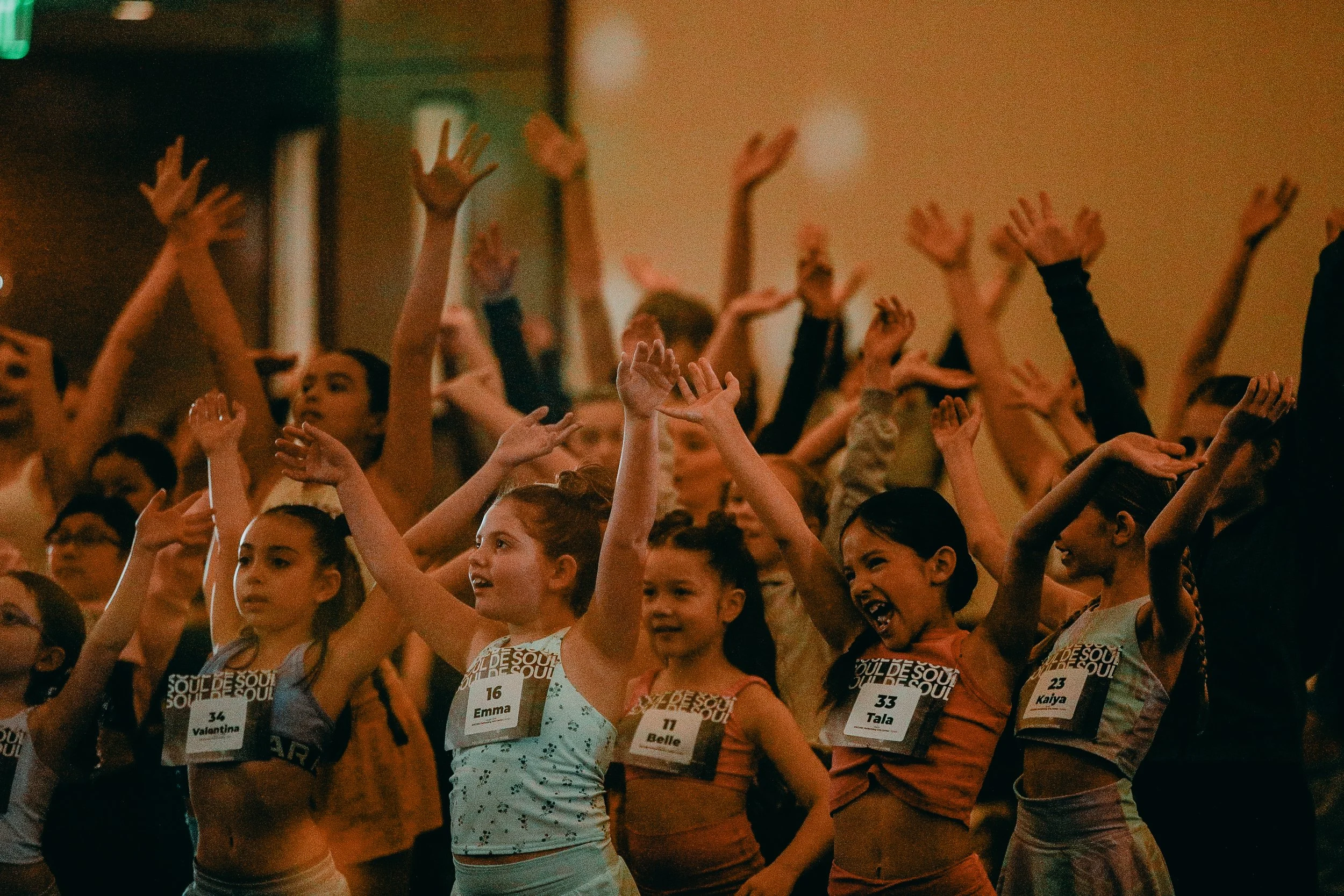 A group of young girls participating in a dance or cheerleading event, raising their hands in the air. They are wearing numbered bibs with names like Valentia, Emma, Belle, Tala, and Kaiya, and appear to be indoors with a warm, ambient lighting.
