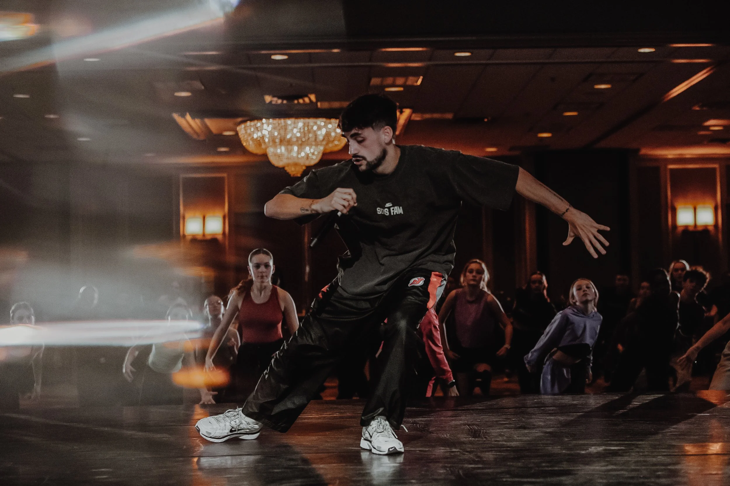 A young woman with dark hair tied up, smiling, wearing a black shirt with white text, in a dimly lit space with bright orange lights and lens flares.