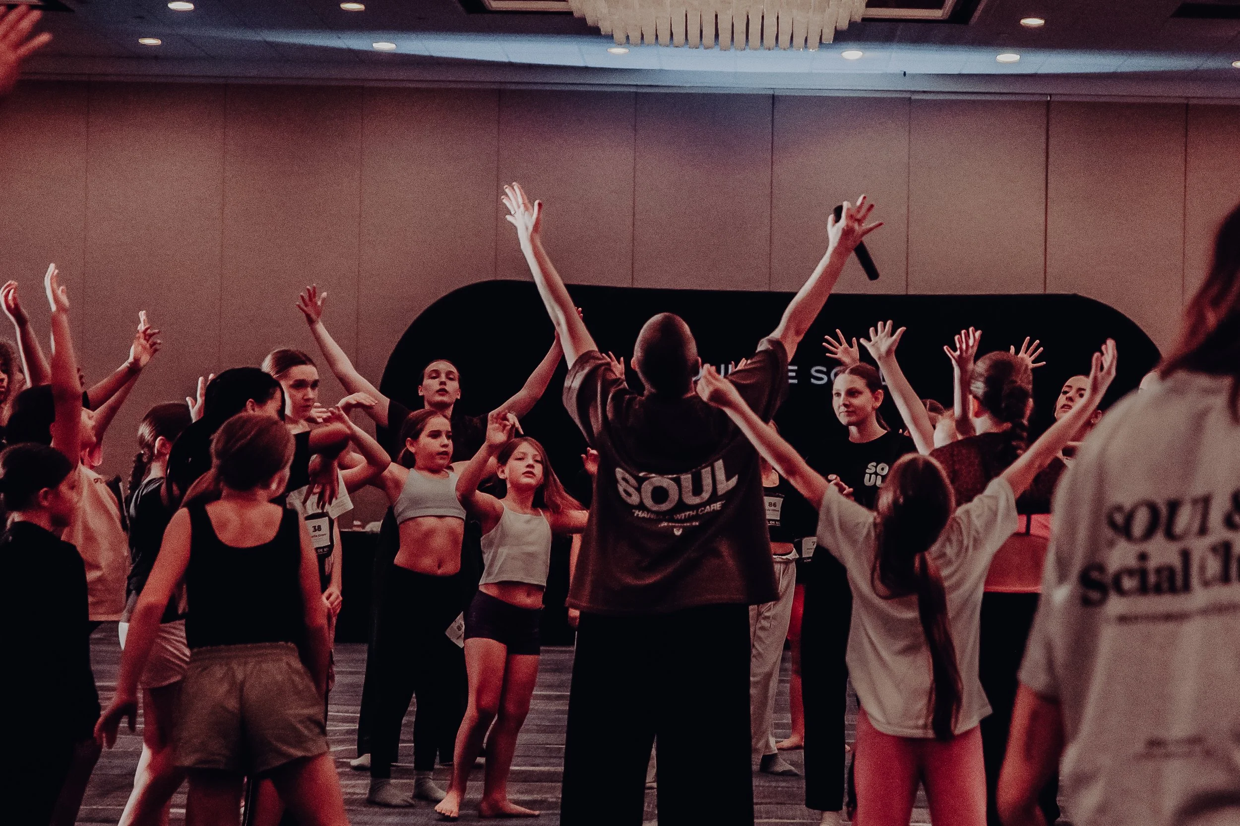 A group of young women and girls participating in a dance or fitness class, raising their arms, in a room with a chandelier and a black backdrop.