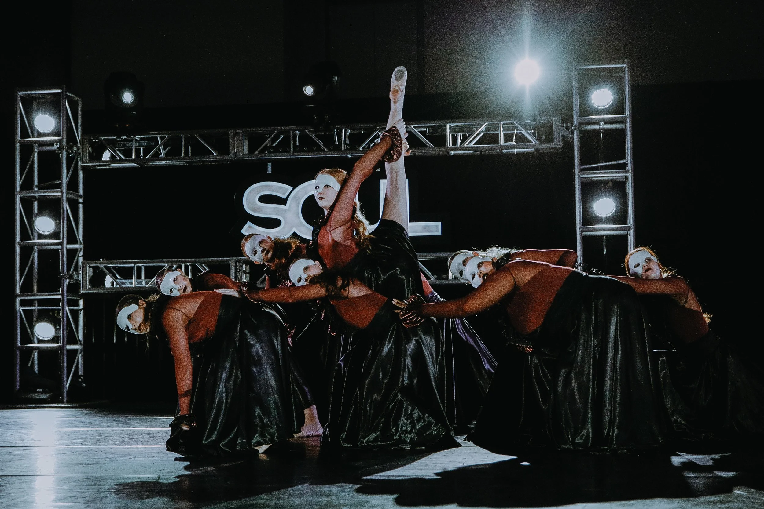 A group of dancers wearing white masks, black skirts, and red tops performing on a stage with metal framework and bright stage lights.