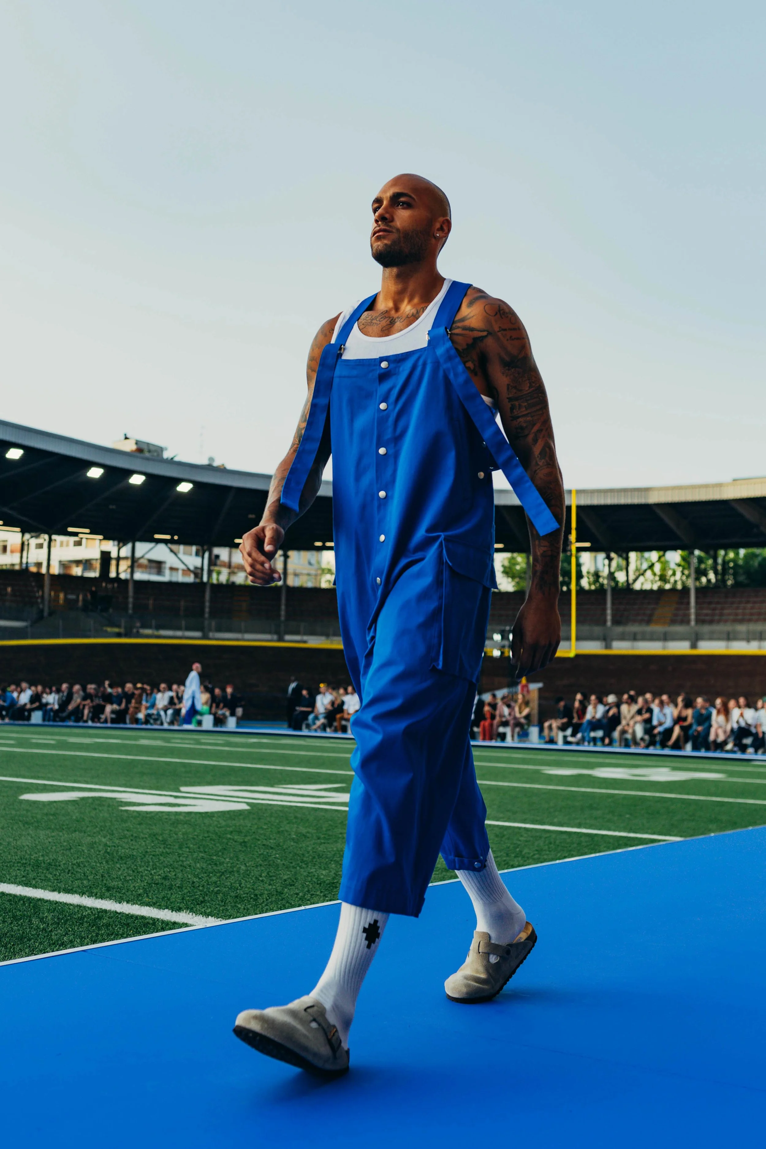 A man with tattoos, wearing a blue jumpsuit and grey slippers, walking on a blue mat on a football field with spectators in the background.