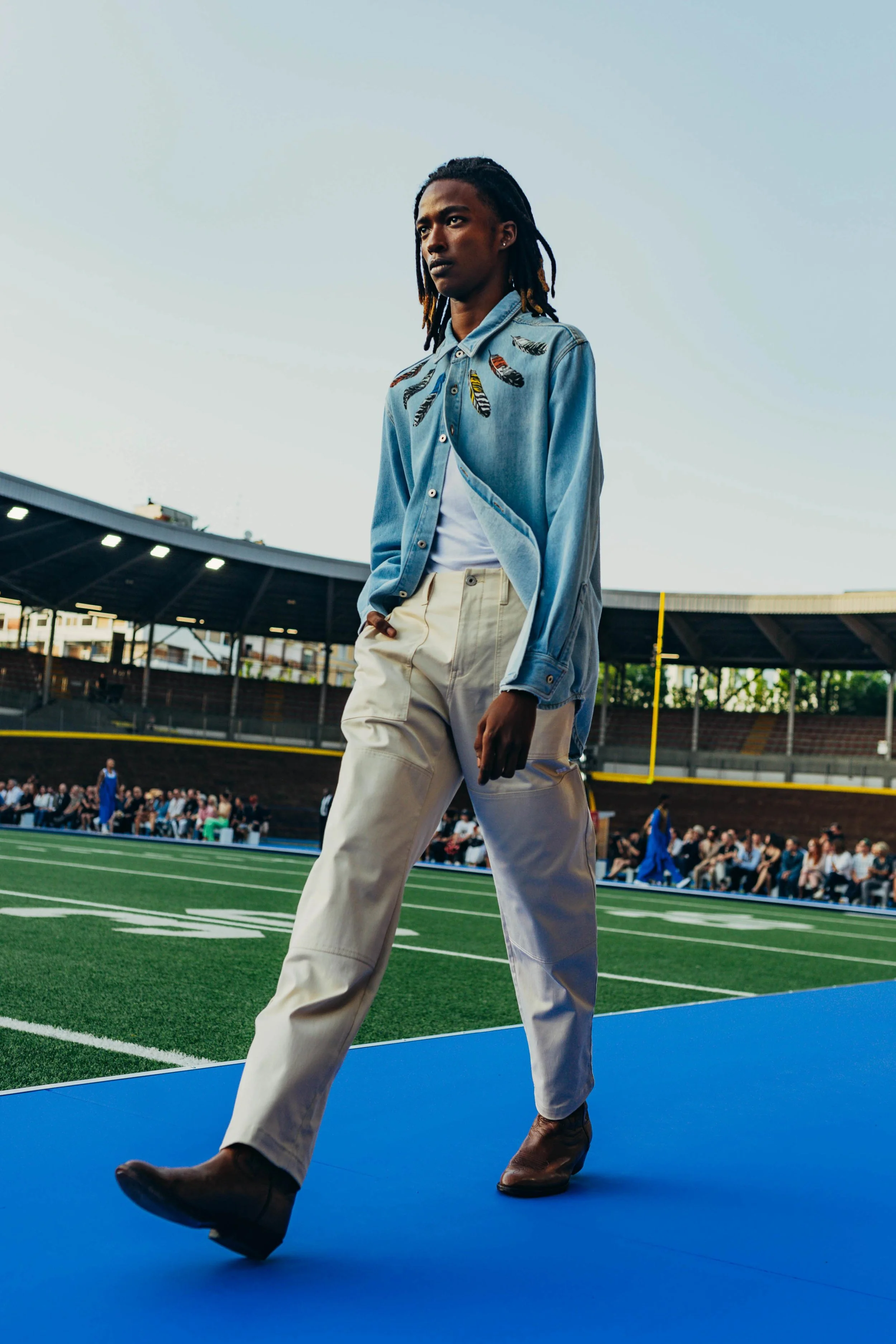 A woman walking on a blue path at a football stadium, with spectators seated in the background, during the daytime.