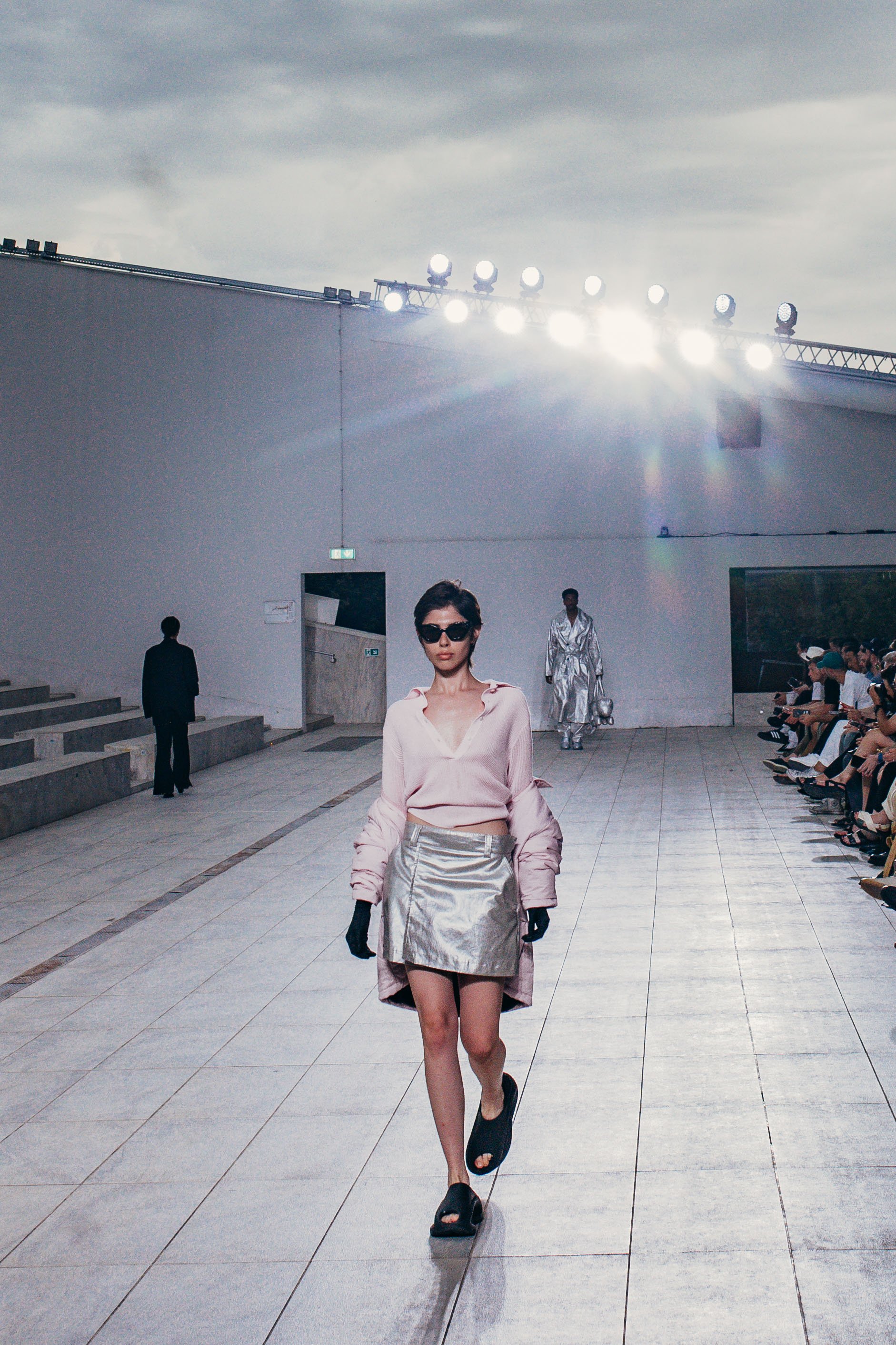 A model walking down a runway at a fashion show, wearing a light pink blouse, metallic silver skirt, black gloves, and black slides, with an audience seated on the right and bright runway lights overhead.
