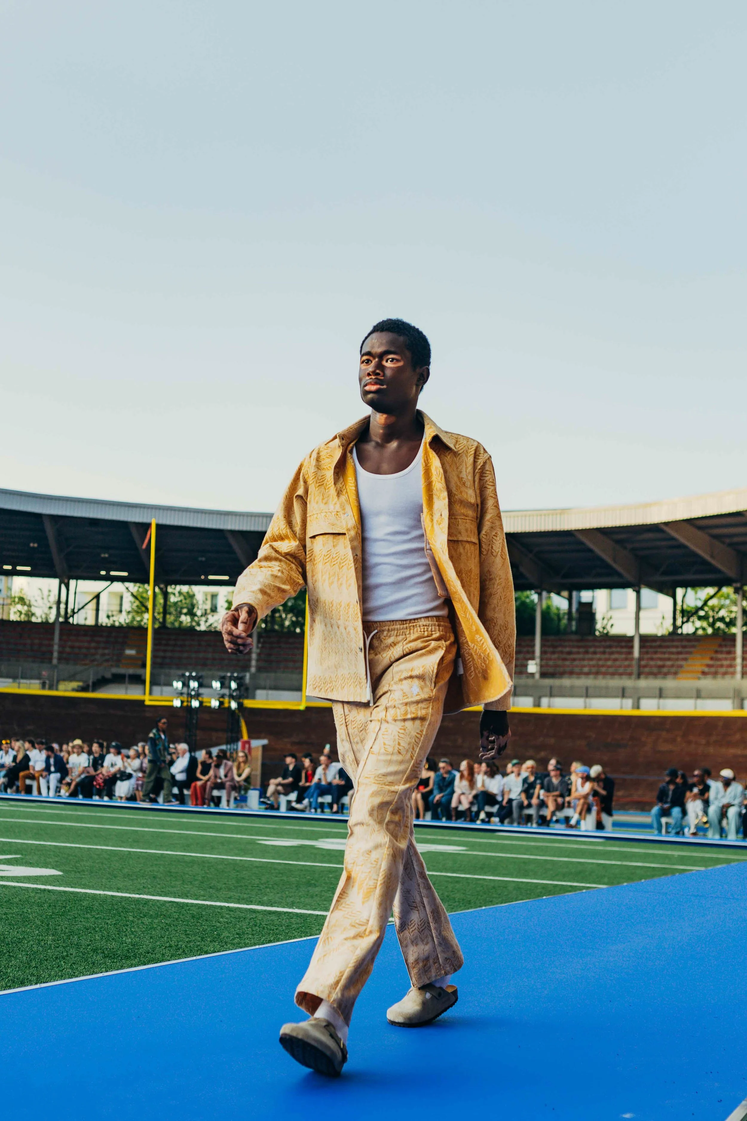 A young man wearing a yellow patterned suit walking on a track at a stadium with a crowd sitting in the background and a goalpost visible.