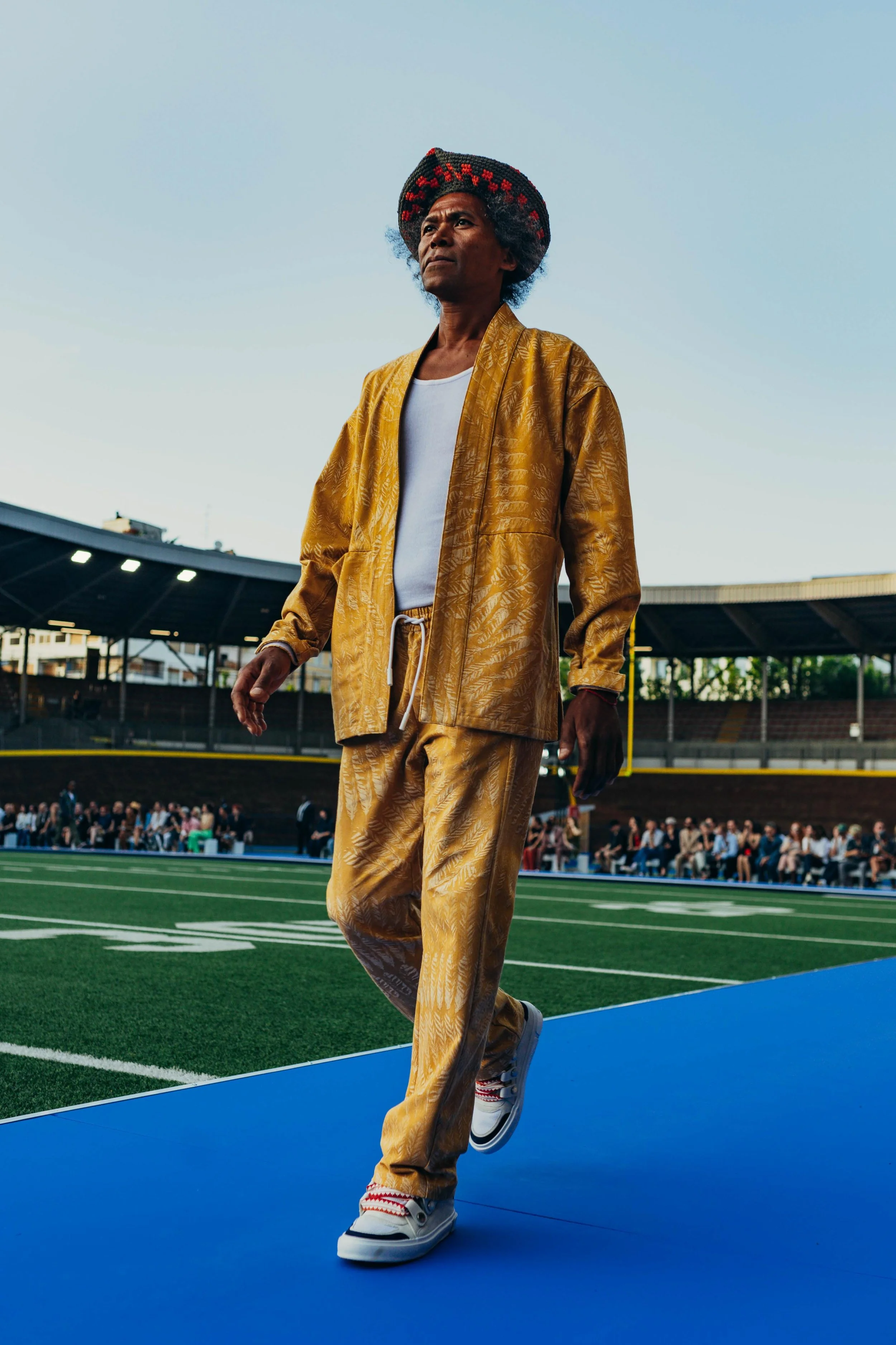 A person walking on a blue track at a sports stadium, wearing a gold-colored patterned jacket and pants, white sneakers, and a hat, with an audience seated in the background.