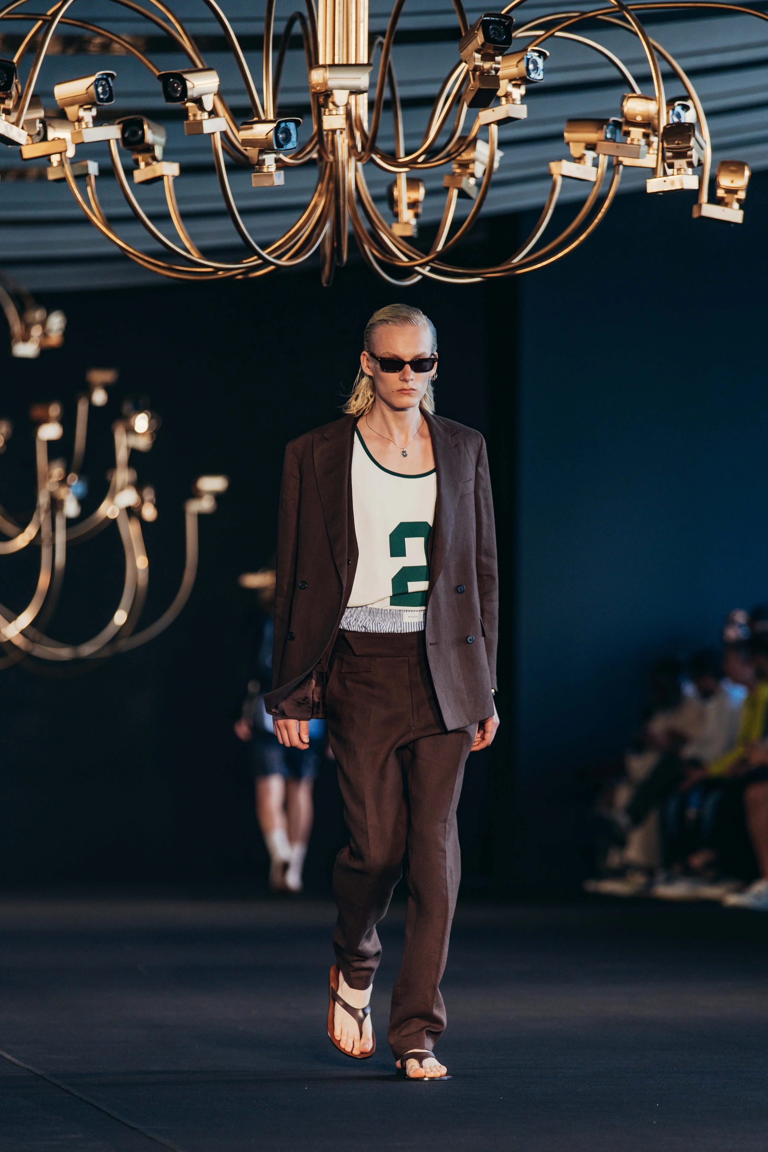 Model walking on runway in fashion show, wearing brown blazer and pants, white top with green number 2, black sunglasses, and flip-flops, under large chandeliers.