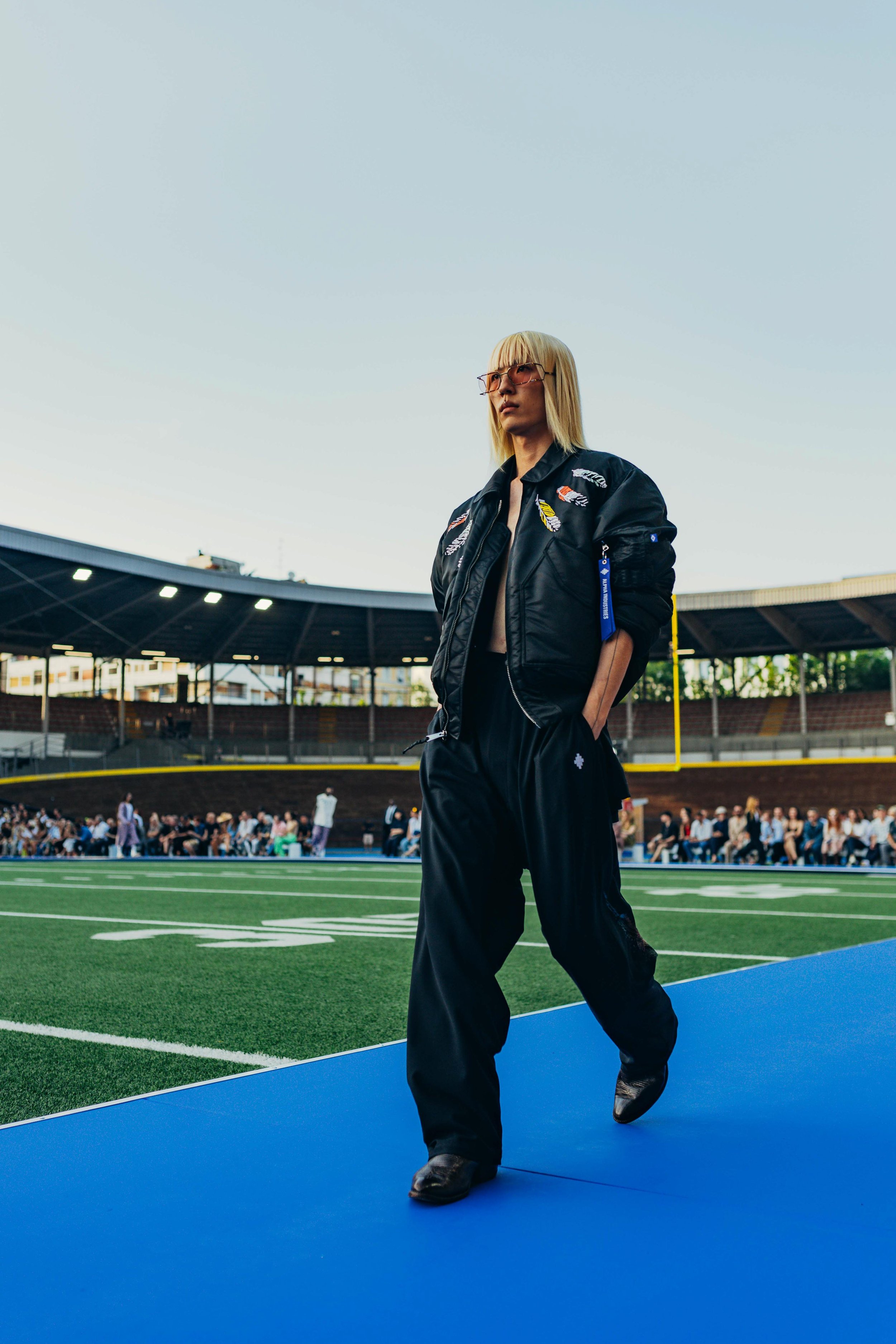 A woman with blonde hair and glasses walking on a blue track in a football stadium, with a crowd in the background.