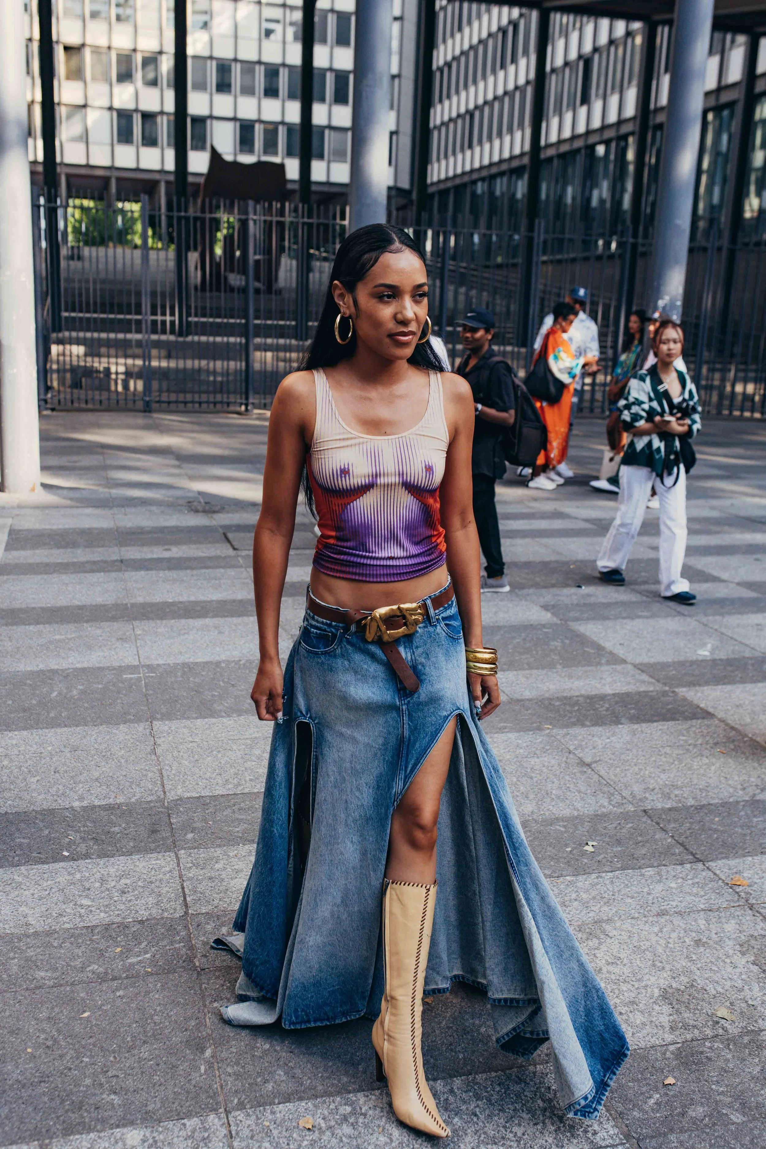 A woman wearing a sleeveless top, wide denim skirt with high slit, and beige high boots standing on a city sidewalk with other pedestrians in the background.