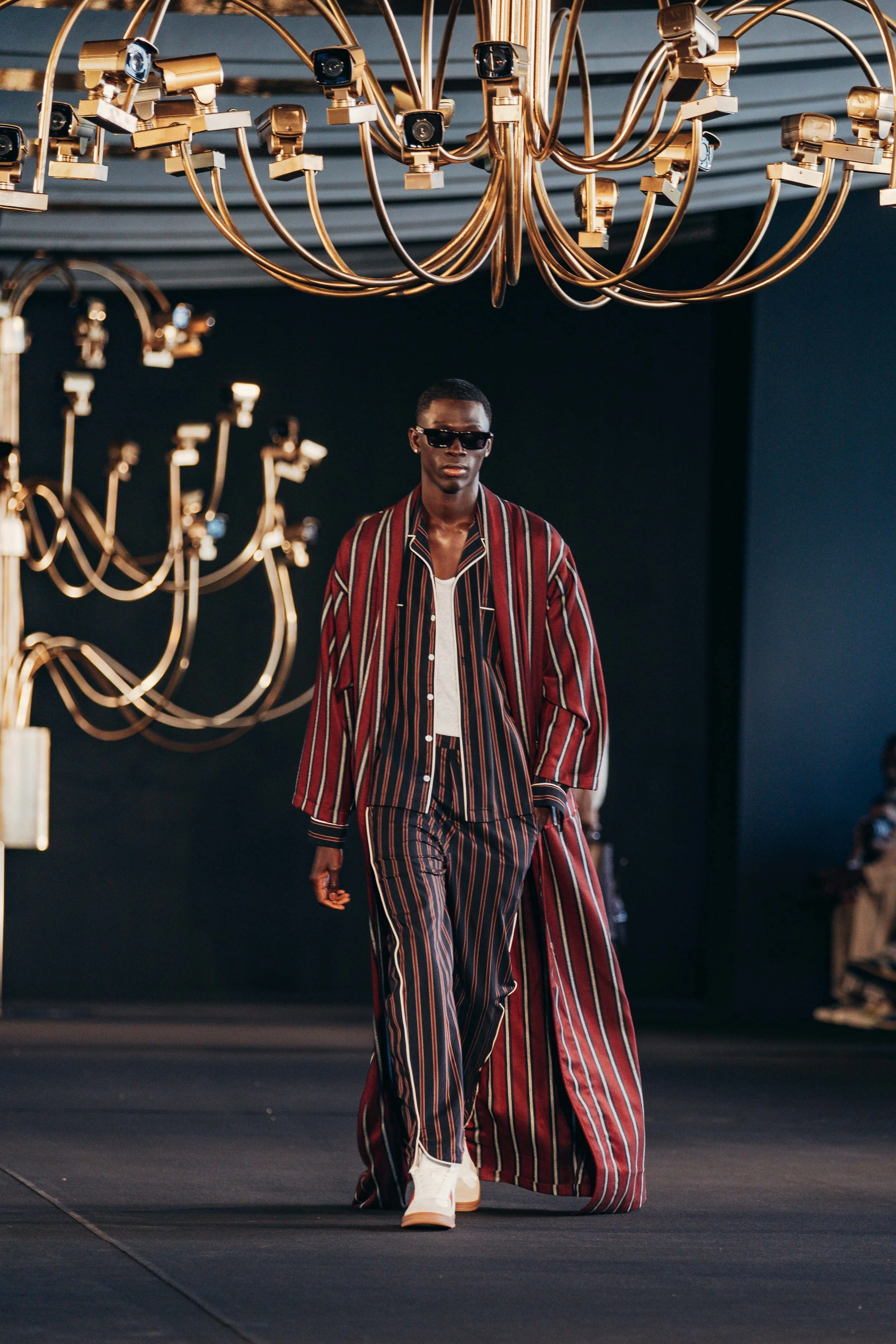 Male model walking on runway wearing striped jacket and pants, sunglasses, and white sneakers, under a large ornate gold chandelier.