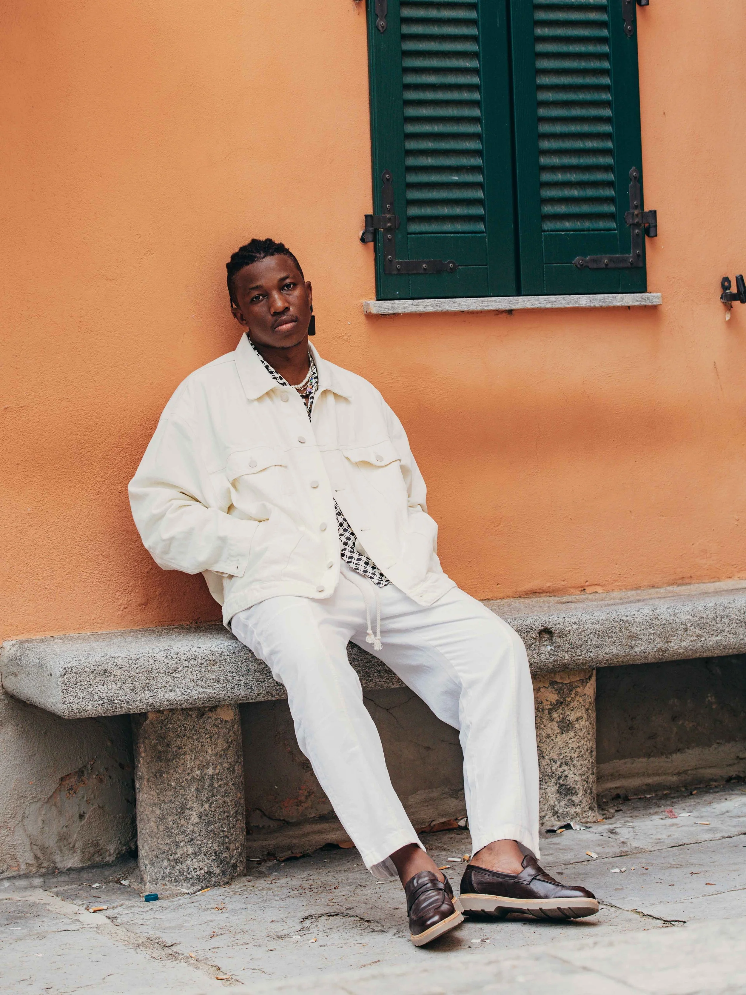 A young man dressed in white clothing sitting on a stone bench with a peach-colored wall and a closed green window behind him.