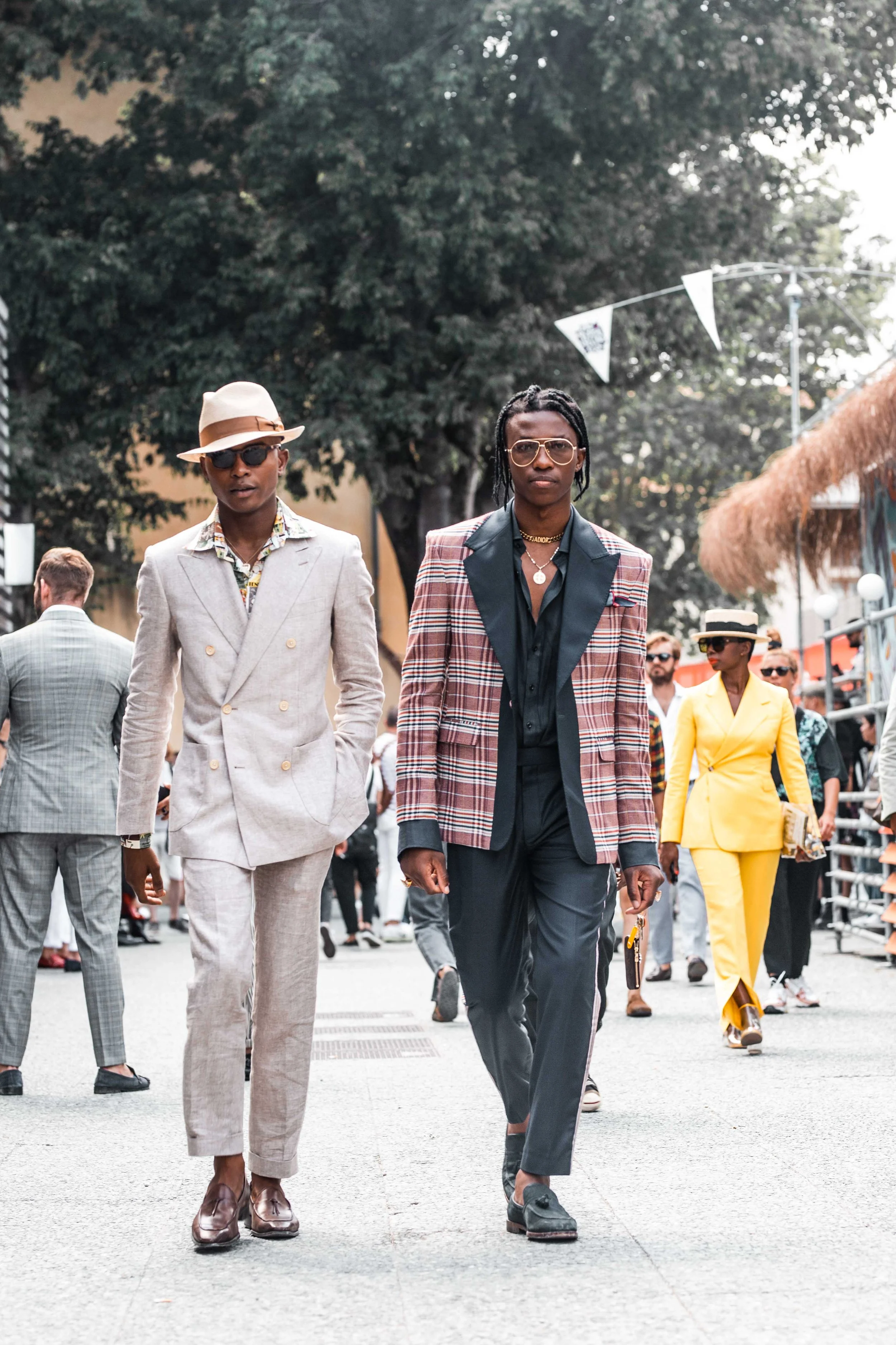 Fashionably dressed people walking outdoors at a street event, with trees and a crowd in the background.