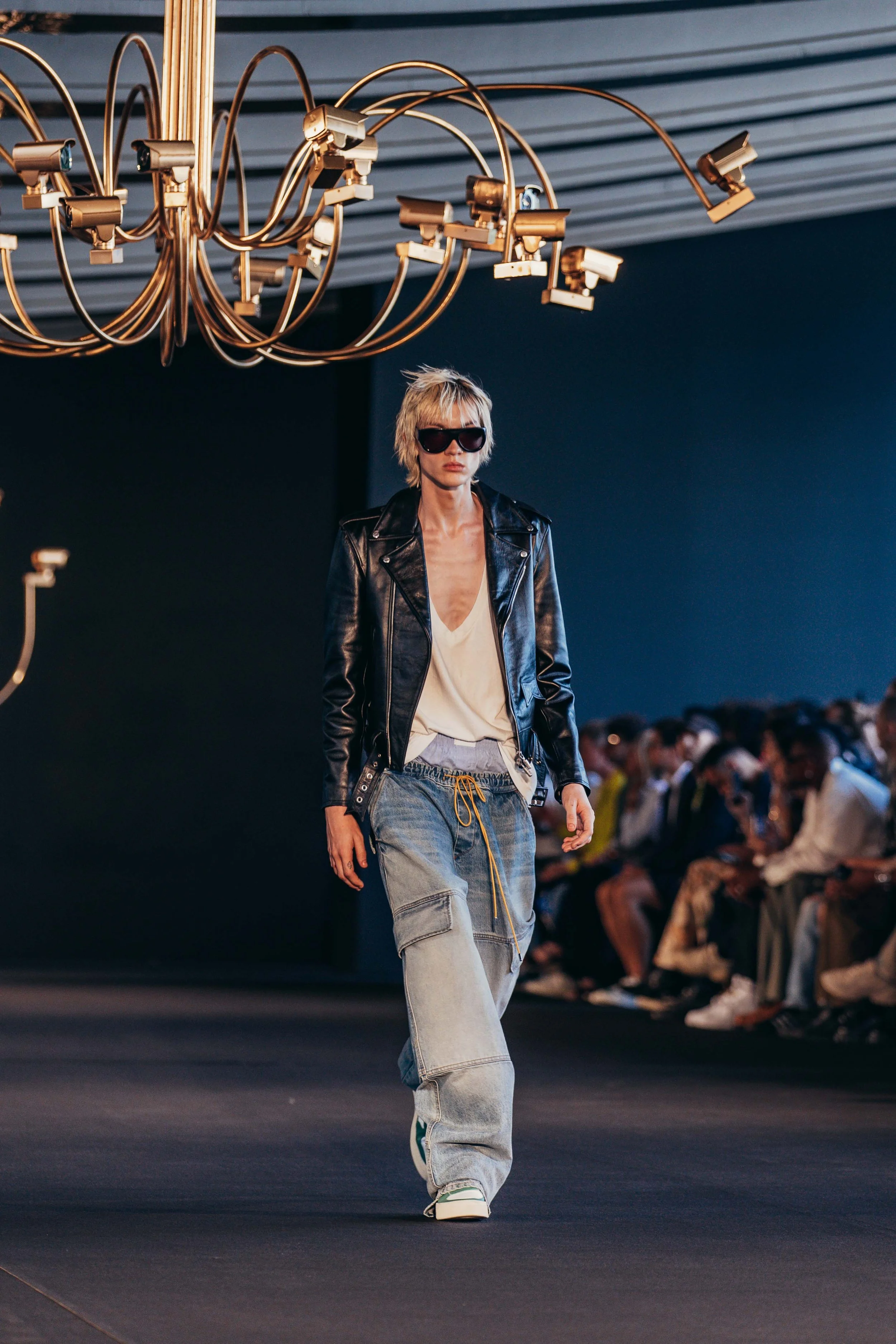Model walking on runway at fashion show, wearing leather jacket, loose cargo jeans, white top, dark sunglasses, under modern chandelier lighting, audience in background.