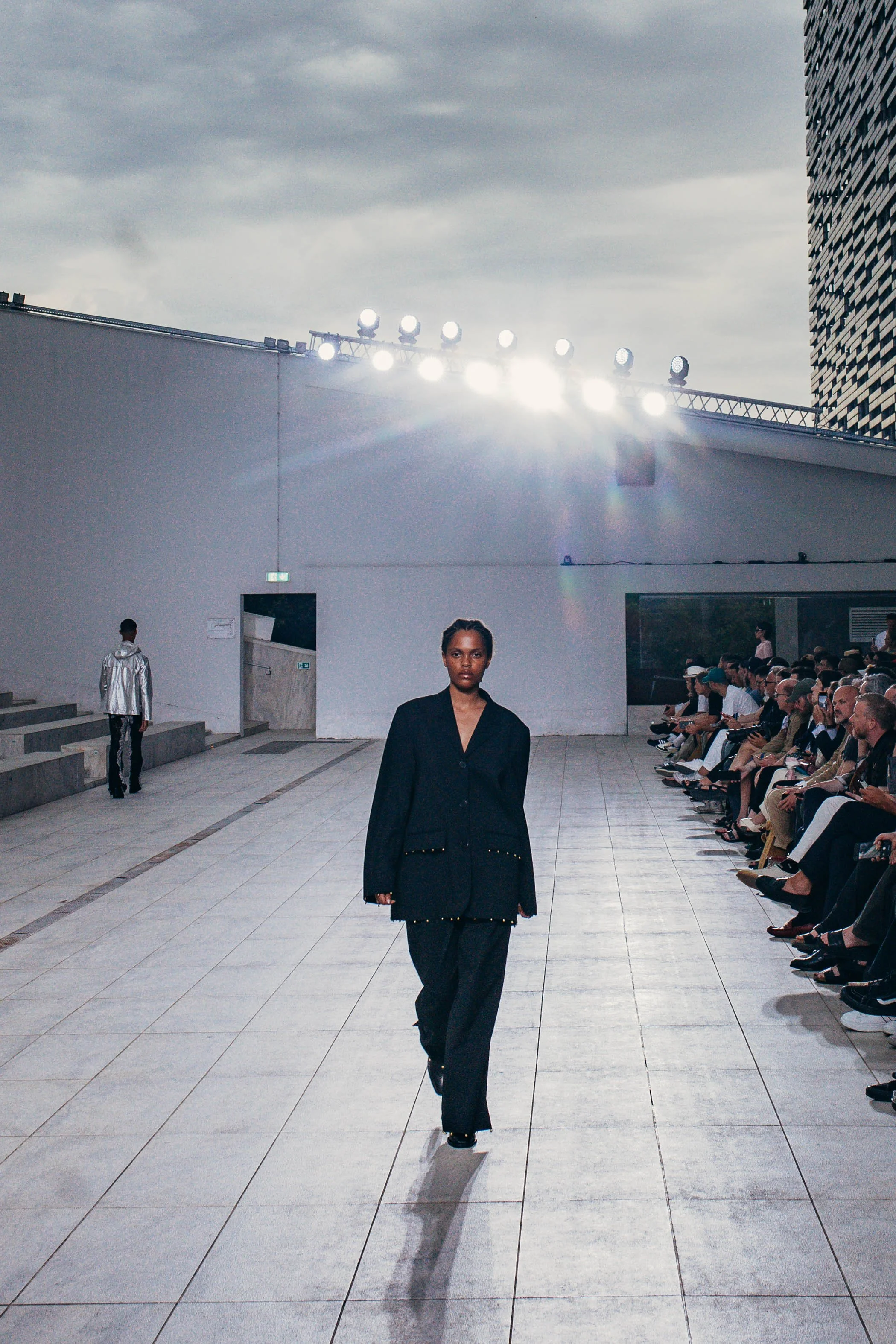 A fashion model walks down an outdoor runway during a fashion show, with audience seated on the right side and a city skyline in the background.