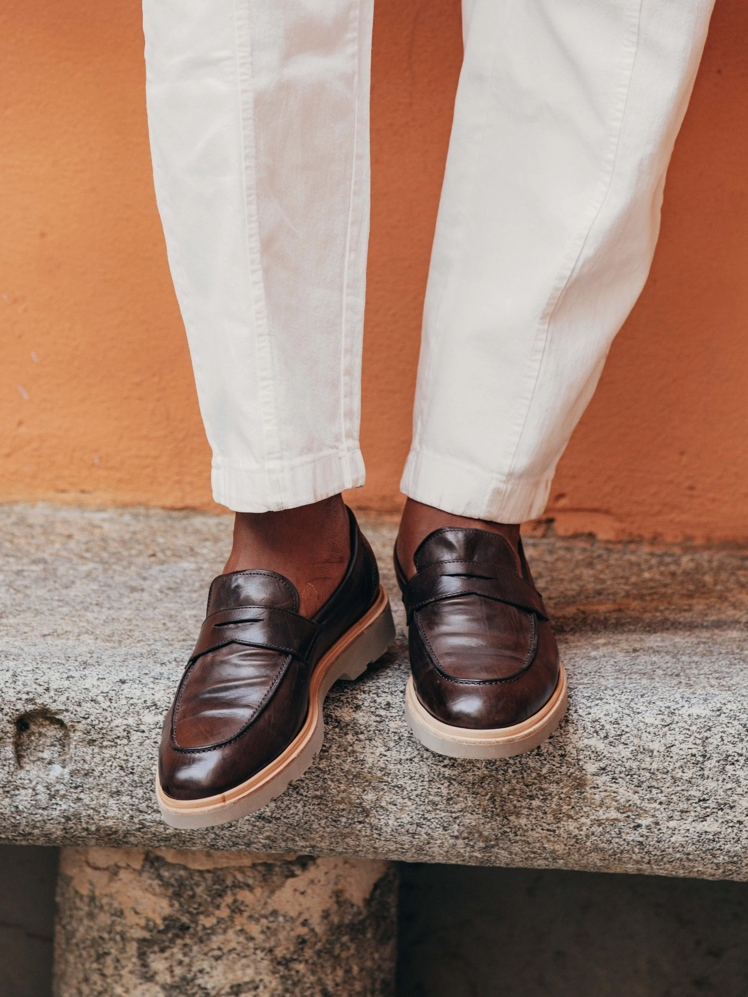 Close-up of a person's feet wearing dark brown leather loafer shoes with beige soles, standing on a stone surface with white pants and an orange wall background.