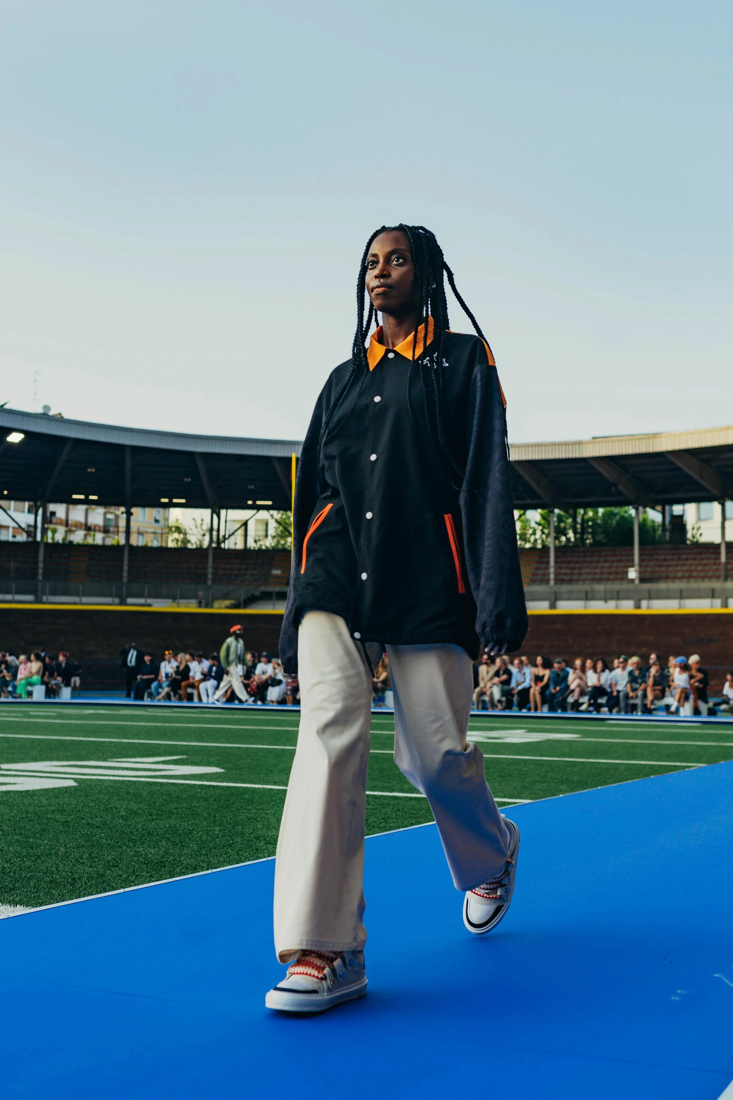 A woman with braided hair walking on a blue track at a sports stadium, with spectators seated in the background during early evening.
