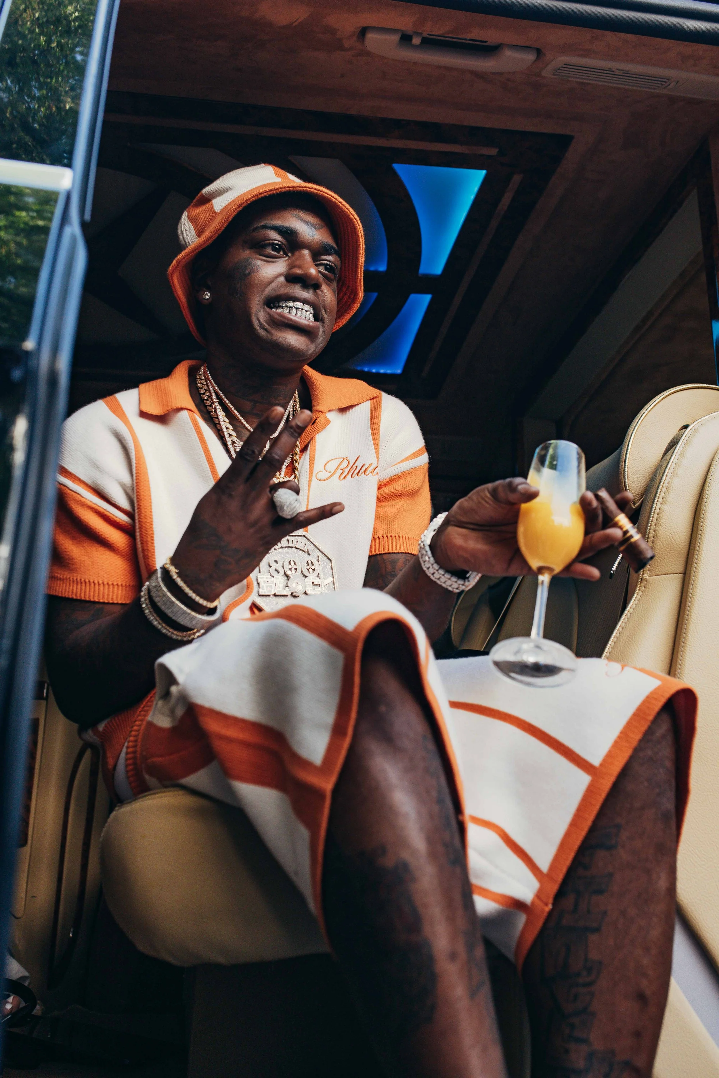 A young man dressed in a white and orange striped outfit and bucket hat sitting inside a luxurious vehicle, holding a cocktail and flashing a peace sign.