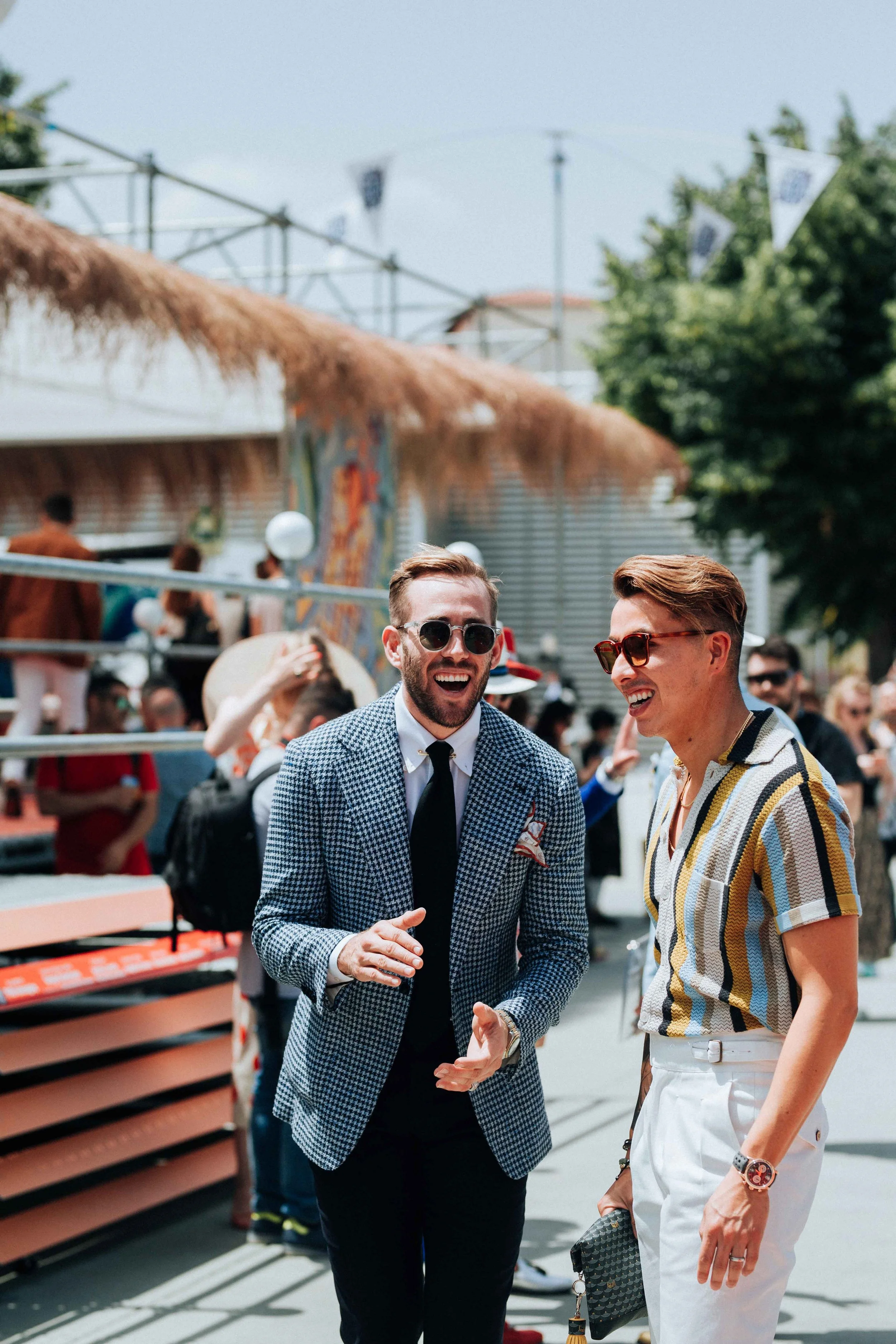 Two men with sunglasses and stylish clothing smiling and talking in a crowded outdoor festival or fair.