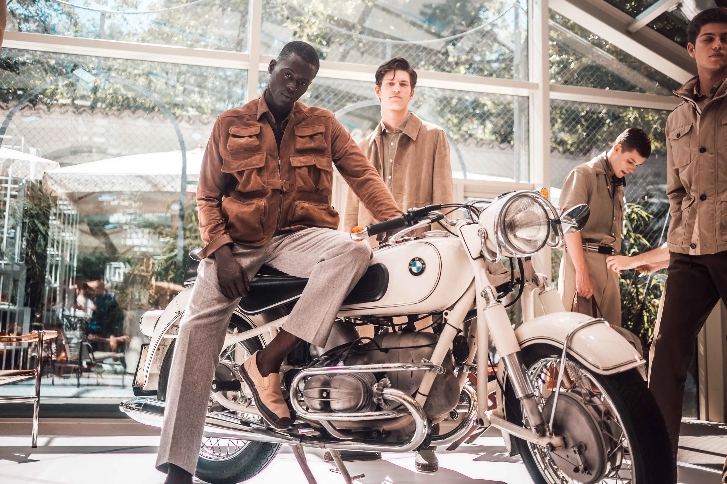 Four young men in stylish clothing are inside a showroom with large windows, with one sitting on a white BMW motorcycle, the other three standing nearby.