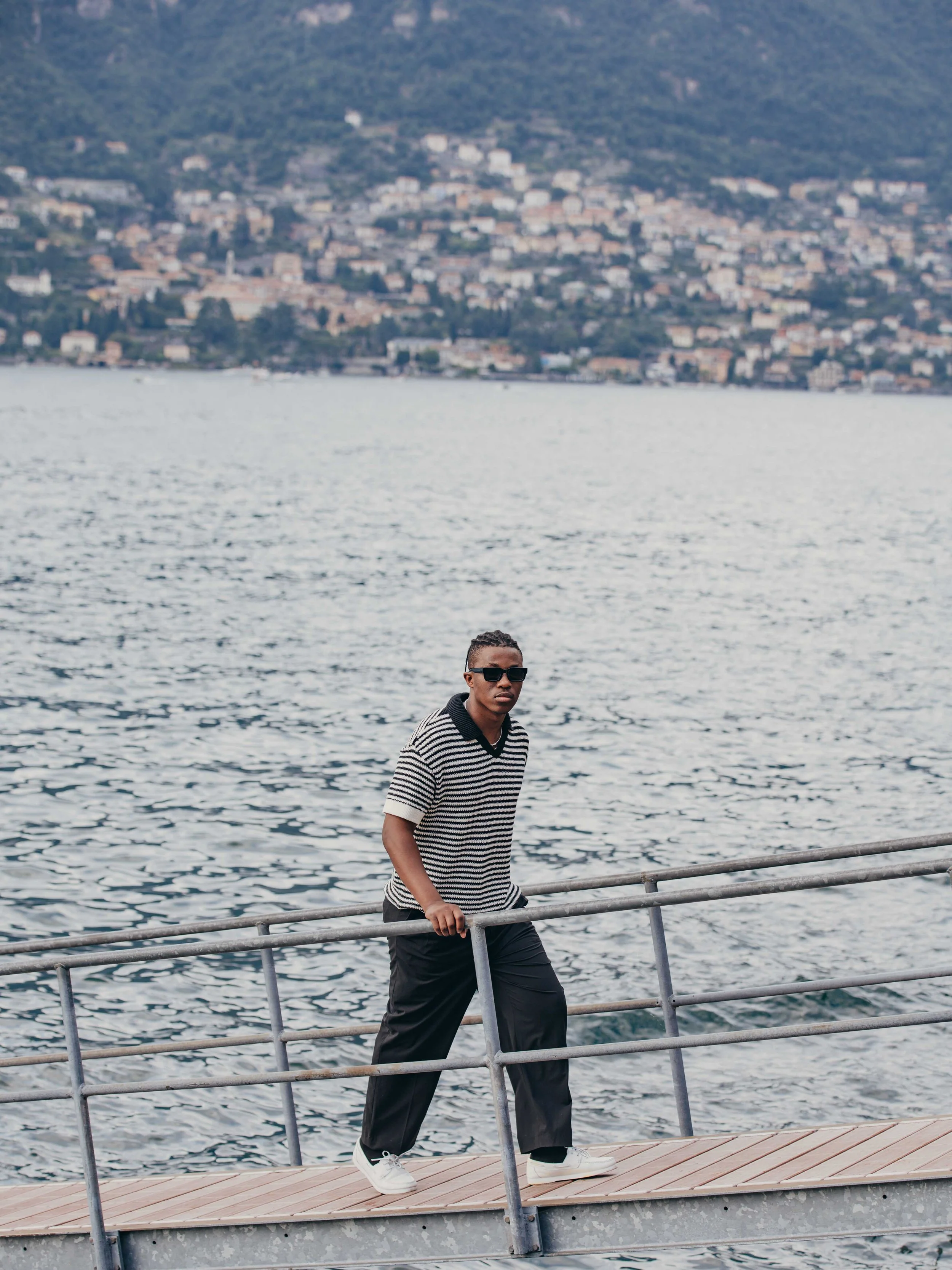 A young man in a striped shirt and black pants walking on a metal dock with water in the background and a cityscape on a hillside.