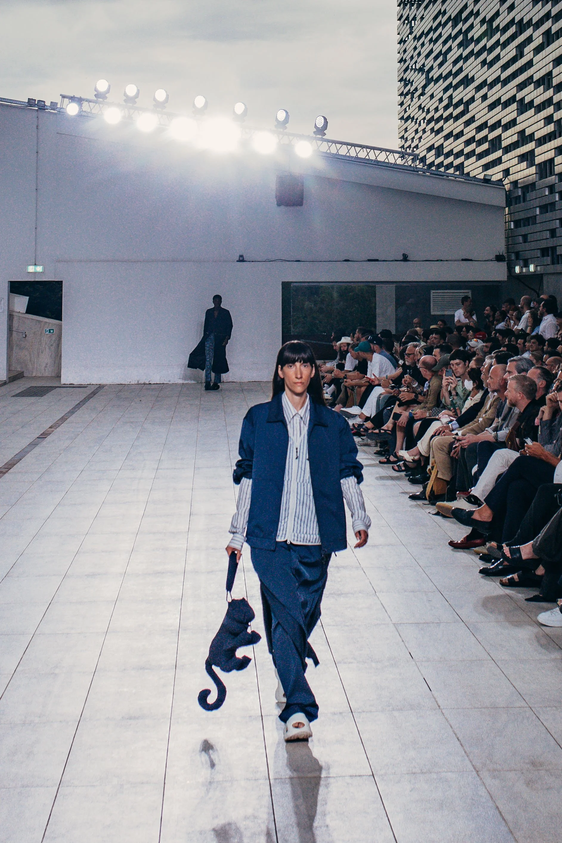 Model walking down runway holding a stuffed black panther at fashion show, audience seated on sides, city skyline in background.