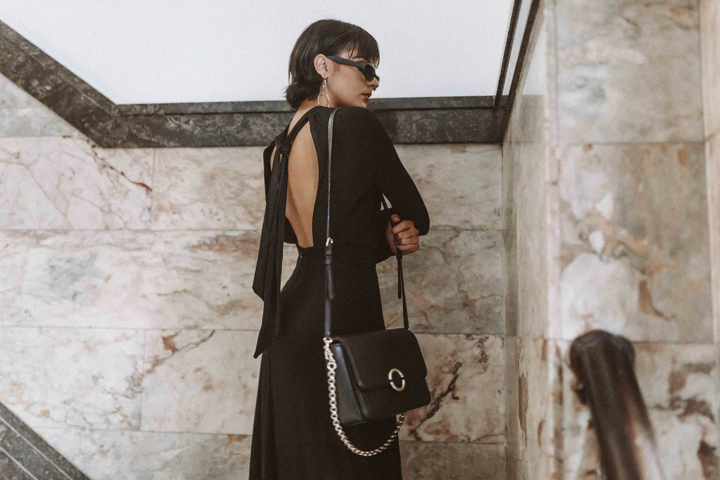 A woman with short dark hair and sunglasses stands on a marble staircase, wearing a backless black dress and holding a black handbag with a gold logo and chain strap.
