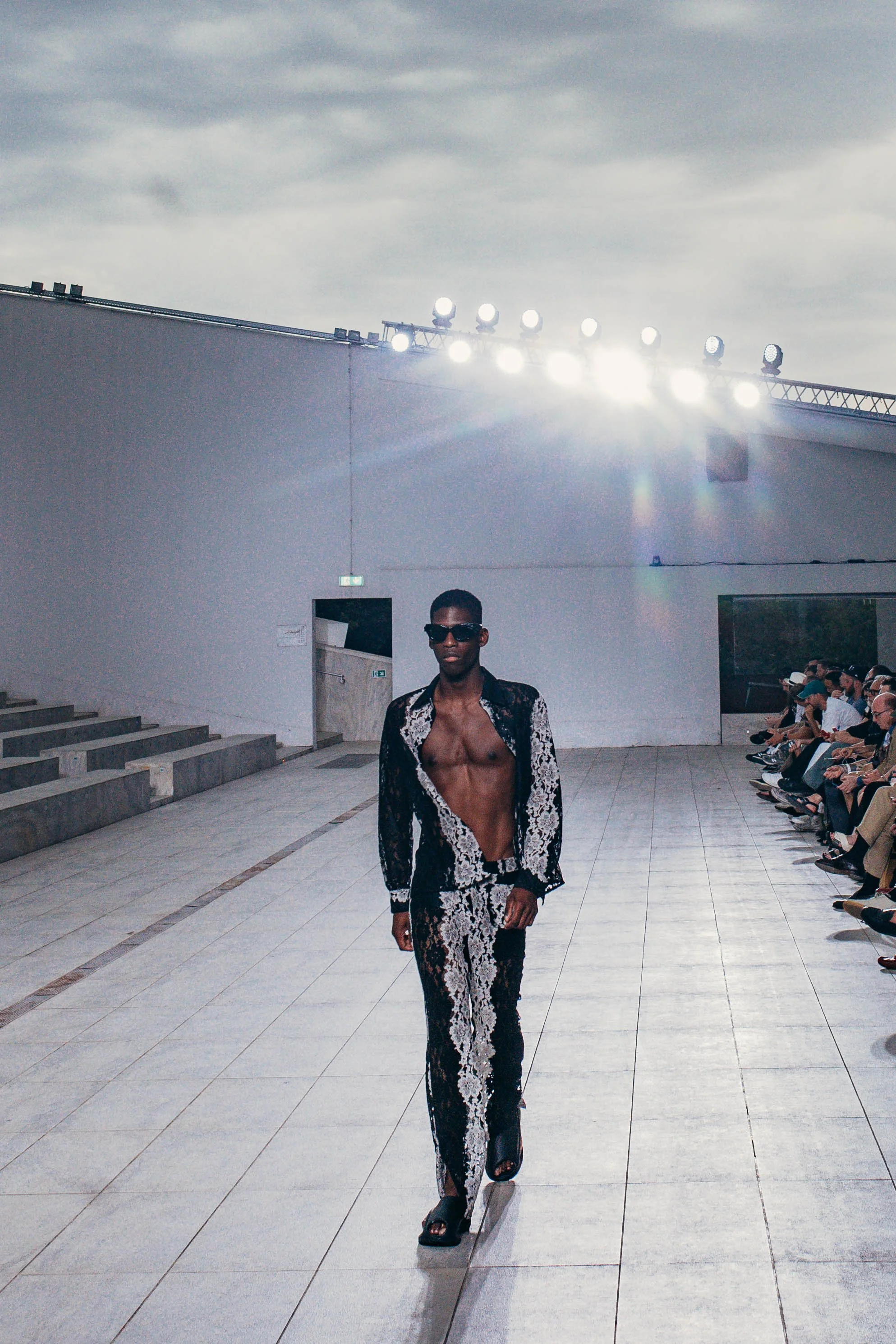 Male model walking on runway wearing black and white floral suit with open shirt, sunglasses, and sandals during fashion show.